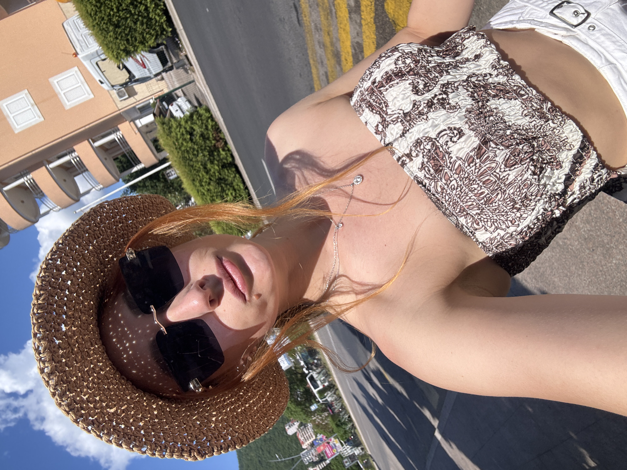 A rotated, close-up selfie captures a young woman outdoors under bright sunshine in Budva, Montenegro. She is wearing a woven straw hat that casts a dappled shadow on her forehead, and large square black sunglasses that conceal her eyes. Her reddish-blonde hair falls over her shoulders and neck. She accessorizes with a delicate silver necklace featuring small charms.
The woman is dressed in a strapless, ruched top with an intricate white and brown paisley-like pattern. Her midriff is visible, and she is wearing white high-waisted shorts or a skirt with a prominent silver buckle. The strong sunlight creates clear shadows on her skin and the surrounding surfaces, indicating a bright, clear day, likely around midday or early afternoon.
The background, viewed from the rotated angle, shows a street scene. On one side, multi-story residential buildings with light peach-colored facades and white balconies are visible, along with parked white vans and green hedges. On the other side, an asphalt road with distinct yellow double lines runs horizontally across the frame. The sky is a vibrant blue with scattered white clouds. The overall scene suggests a sunny, relaxed vacation setting. No text is visible in the image.