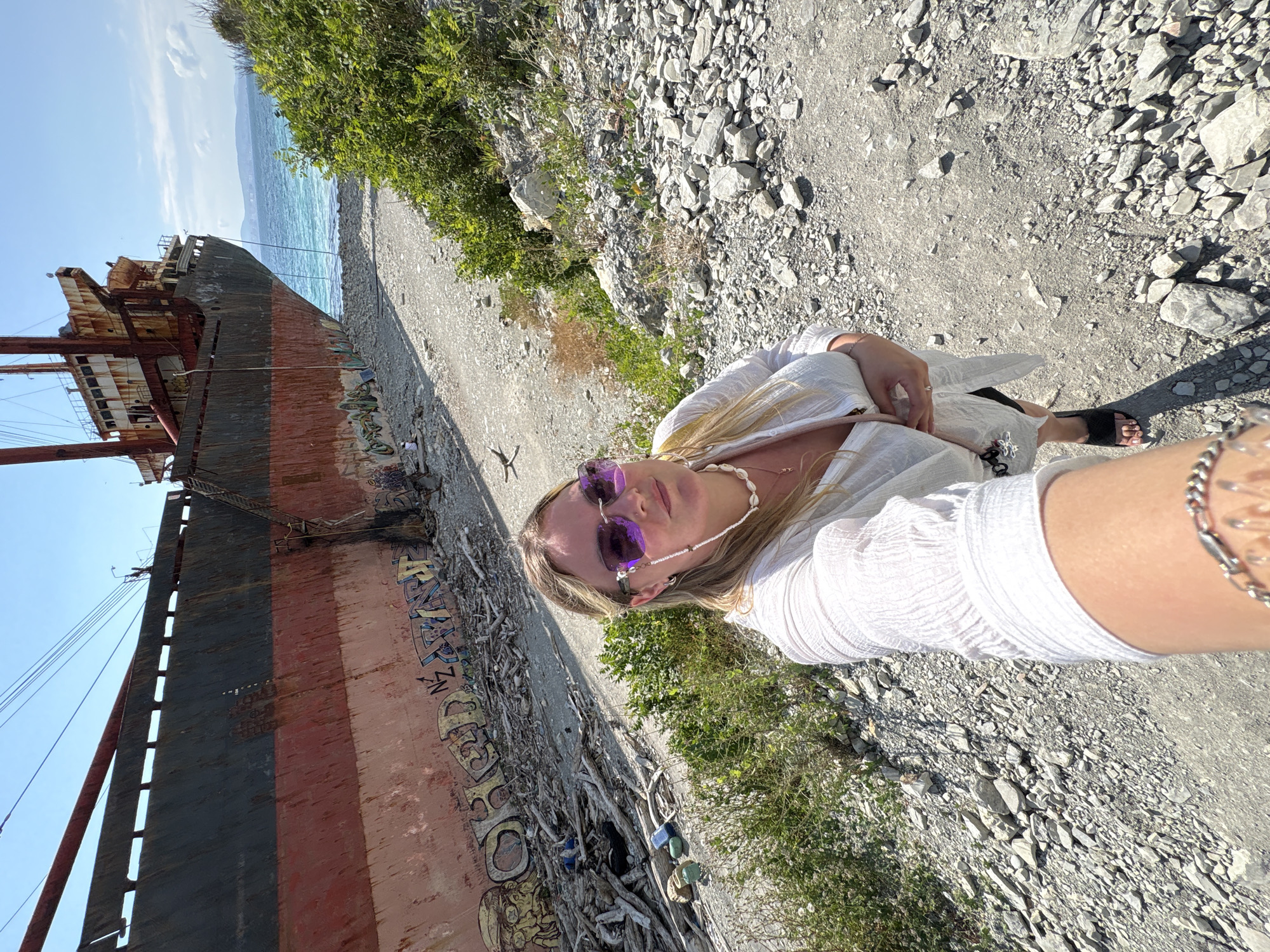A woman with blonde hair wearing sunglasses and a white top poses for a selfie in front of a beached, rusted cargo ship in Kabardinka, Russia. The ship is partially submerged and covered in graffiti. The foreground is a rocky, barren ground with some green bushes. The background shows the sea and distant hills under a clear blue sky.