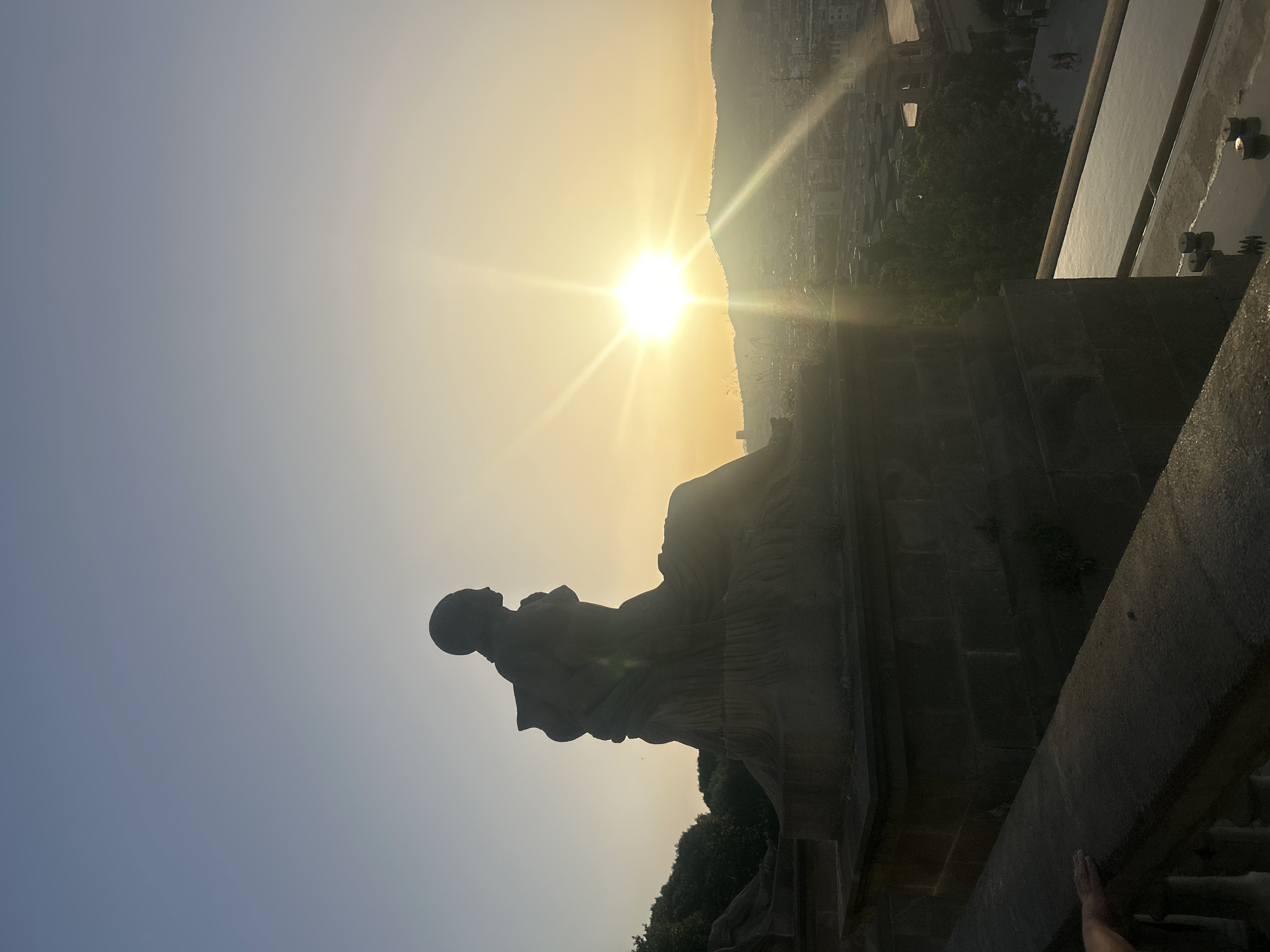 This is a photograph taken at sunset from a vantage point overlooking Barcelona, Spain. In the foreground, a large stone sculpture of a seated figure is silhouetted against the bright sky. The sculpture is positioned on a stone platform. To the right of the sculpture, the sun is setting, casting a warm glow and creating visible light rays. Beyond the sculpture, the city of Barcelona stretches out, with buildings and a hill topped by a structure visible in the distance. The sky transitions from a clear blue at the top to a golden hue near the horizon. In the lower right portion of the image, a shallow pool of water and several small circular objects are visible. The lower left shows a balustrade with a hand resting on it, suggesting a person is present.