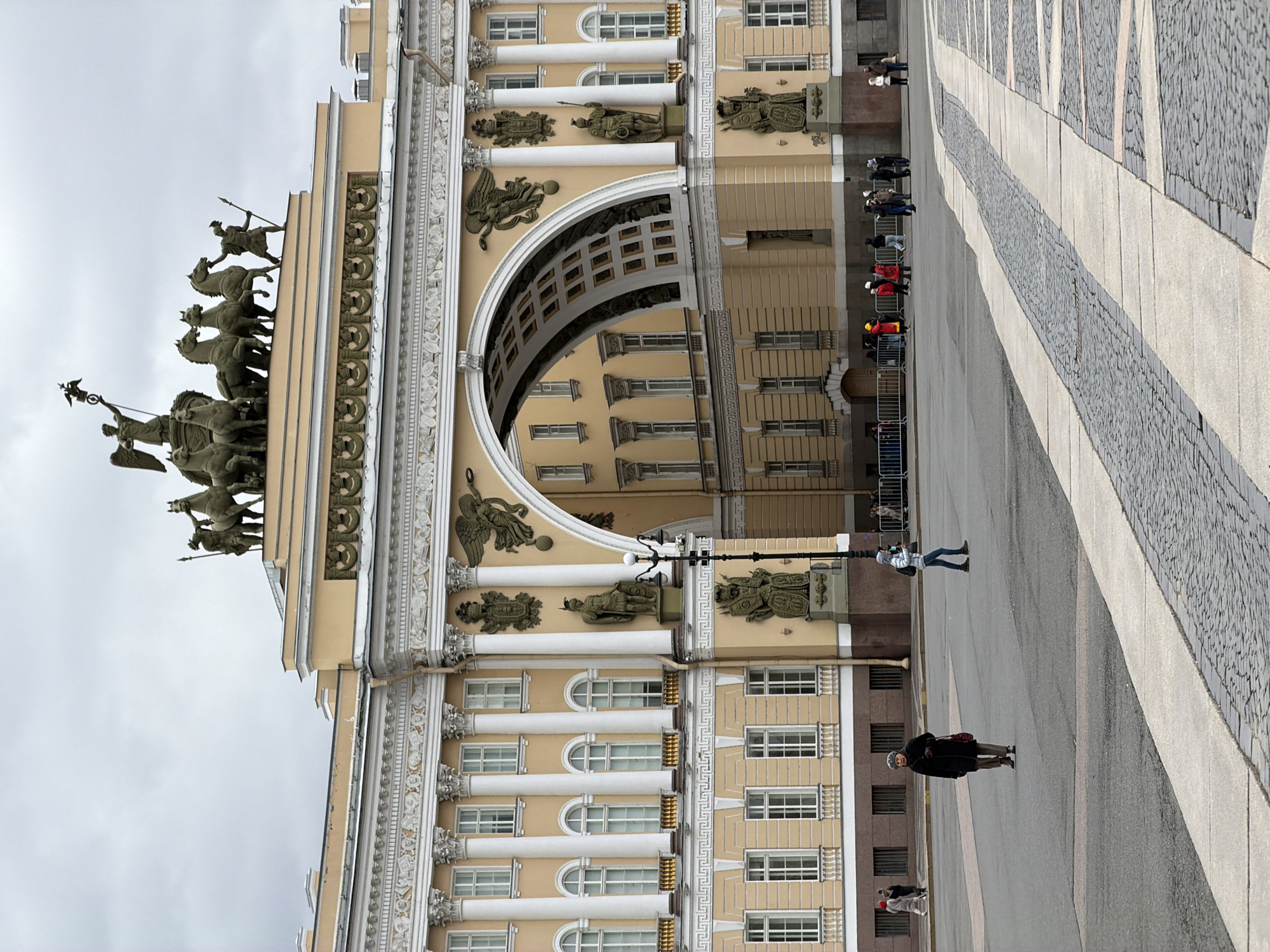 The image captures a broad view of the General Staff Building on Palace Square in Saint Petersburg, Russia, under an overcast sky. The grand, neoclassical building, painted in a light yellow/beige hue with white architectural detailing, features a prominent triumphal arch dominating the upper facade.
Perched atop the arch is the monumental bronze sculpture group known as the Chariot of Glory (or Quadriga), depicting a winged goddess, Victoria, driving a six-horse chariot with a soldier figure leading the horses. The building's facade is richly adorned with white columns, pilasters, decorative cornices, intricate bas-reliefs featuring warrior figures, and statues. Numerous windows are visible across the building's multiple levels.
In the foreground and extending across the right side of the frame is the wide expanse of Palace Square, paved with a pattern of light-colored flagstones and darker cobblestones, forming distinct pathways. Several individuals are present in the square. One person in a dark coat and hat is walking in the mid-ground. Further back, a group of people is standing behind white metal crowd barriers, possibly queuing or observing.
The overall scene depicts a quiet daytime moment in a historically significant urban setting. The lighting is soft and diffused due to the cloudy weather, casting no harsh shadows. No specific event beyond general public presence is evident, and no discernible text appears in the frame.