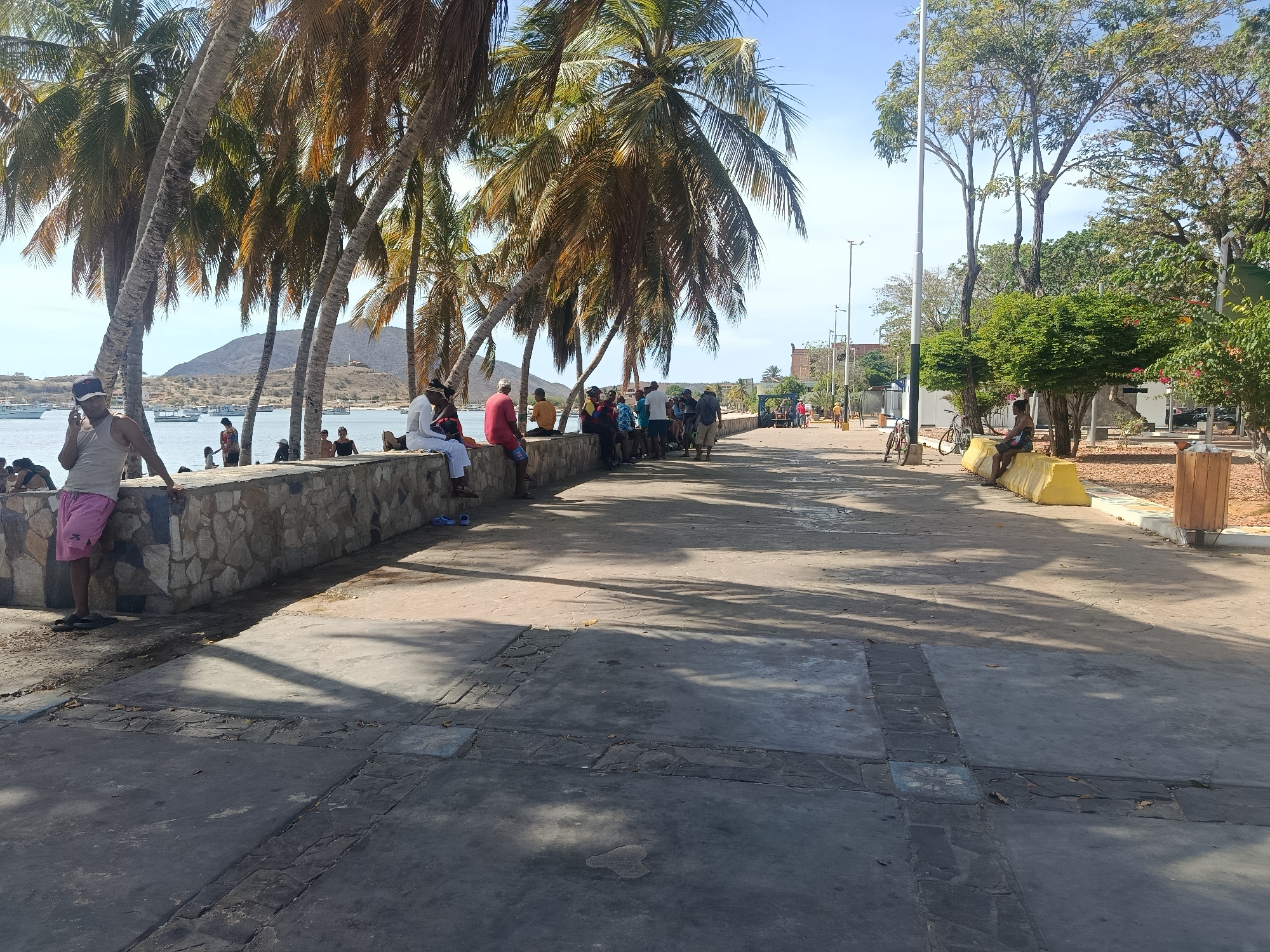 This is an outdoor scene in Juan Griego, Venezuela. The foreground features a wide concrete walkway with sections of darker and lighter gray stones. Palm trees line the left side, casting shadows on the walkway. Along the seawall to the left, several people are seated or standing. A man with a hat and tank top stands at the edge of the wall near the bottom-left corner, seemingly using his phone. Others are gathered in groups, some wearing brightly colored clothing. In the background, a bay with boats and a distant hill are visible. To the right of the walkway is a park-like area with trees, benches, and lampposts. A person is sitting on a yellow barrier along the walkway. In the far distance, buildings and other people can be seen. The sky is light blue with some clouds.
