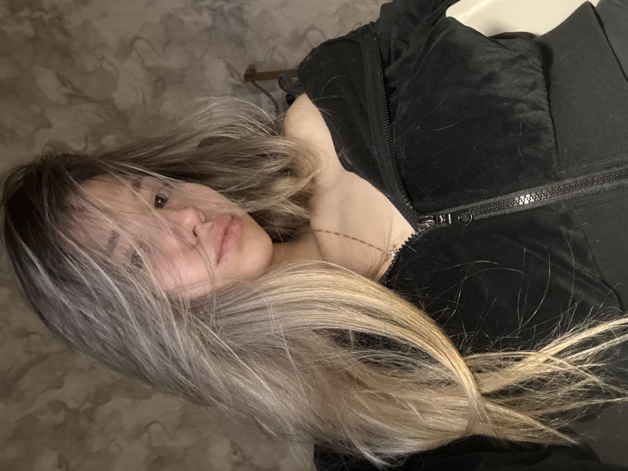 A young woman with long, ombre hair is lying down, facing the camera. Her hair transitions from dark roots to blonde ends, and it partially covers her face. She is wearing a dark, possibly velvet, zip-up jacket. The background is a mottled pattern, likely a wall or other surface. No location information regarding Noviy, Russia is present in the image.