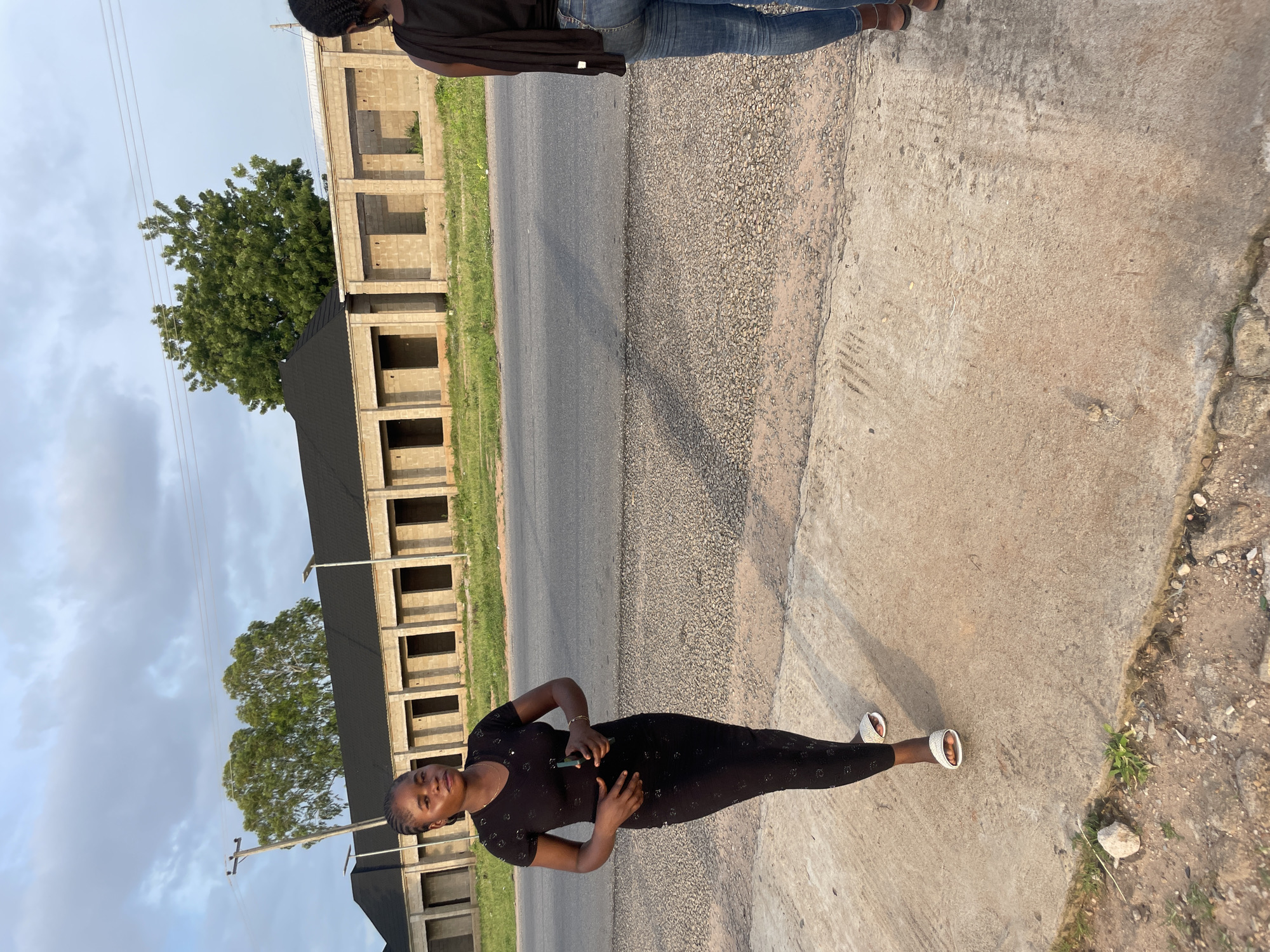 A young Black woman stands on a concrete sidewalk, facing forward and holding a dark object in her right hand. She is wearing a black, form-fitting, short-sleeved dress with a patterned texture, and white slide sandals. To her right, another Black woman, seen from the back, is wearing blue jeans and a black top, with a dark scarf draped over her shoulders.
They are positioned on a sidewalk adjacent to a road with a gravel shoulder. In the background, across the road, is a row of unfinished, two-story buildings constructed with concrete blocks, topped with dark roofs. Green grass and a few trees are visible around the buildings. The sky above is partly cloudy. The scene appears to be a street in Jalingo, Nigeria, possibly a developing area with ongoing construction. The lighting suggests it is late afternoon or early evening, with long shadows cast on the ground.
