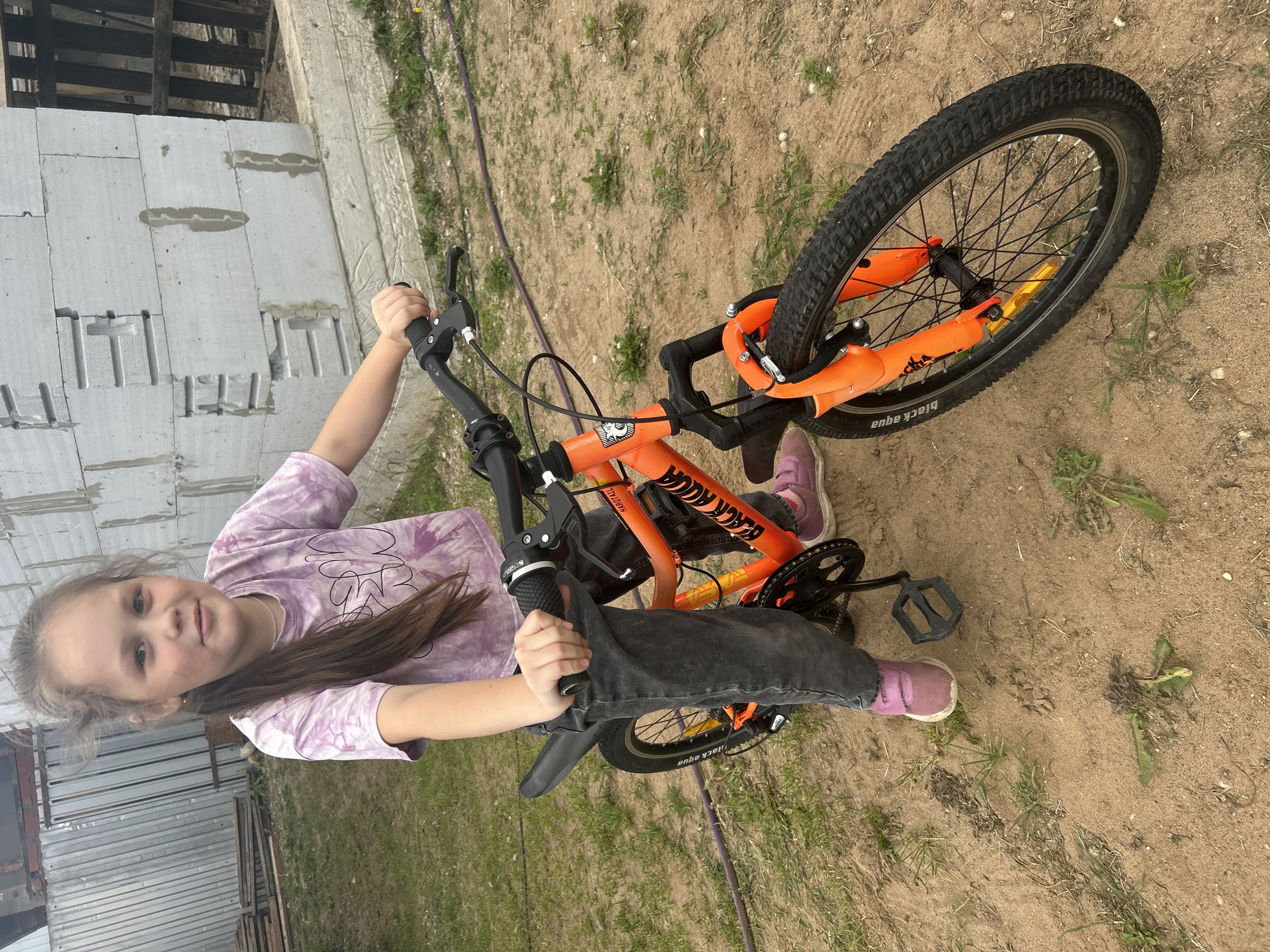 An outdoor full shot shows a young Caucasian girl with long brown hair and light skin, wearing a purple tie-dye t-shirt and dark gray jeans, posing with a bright orange bicycle. The girl is seated on the bicycle, with her right hand on the handlebar. She is looking directly at the camera with a slight smile. The bicycle is a 20-inch model, with black tires and black rims, and orange handlebars and frame. The background consists of a sandy ground with sparse green grass, a metal fence on the left, and a partially constructed building made of gray concrete blocks on the right. A purple hose lies on the ground in the mid-ground. The scene appears to be in a residential or construction area in the city of Chaikovsky, Russia, based on the presence of the uncompleted structure and the general environment.