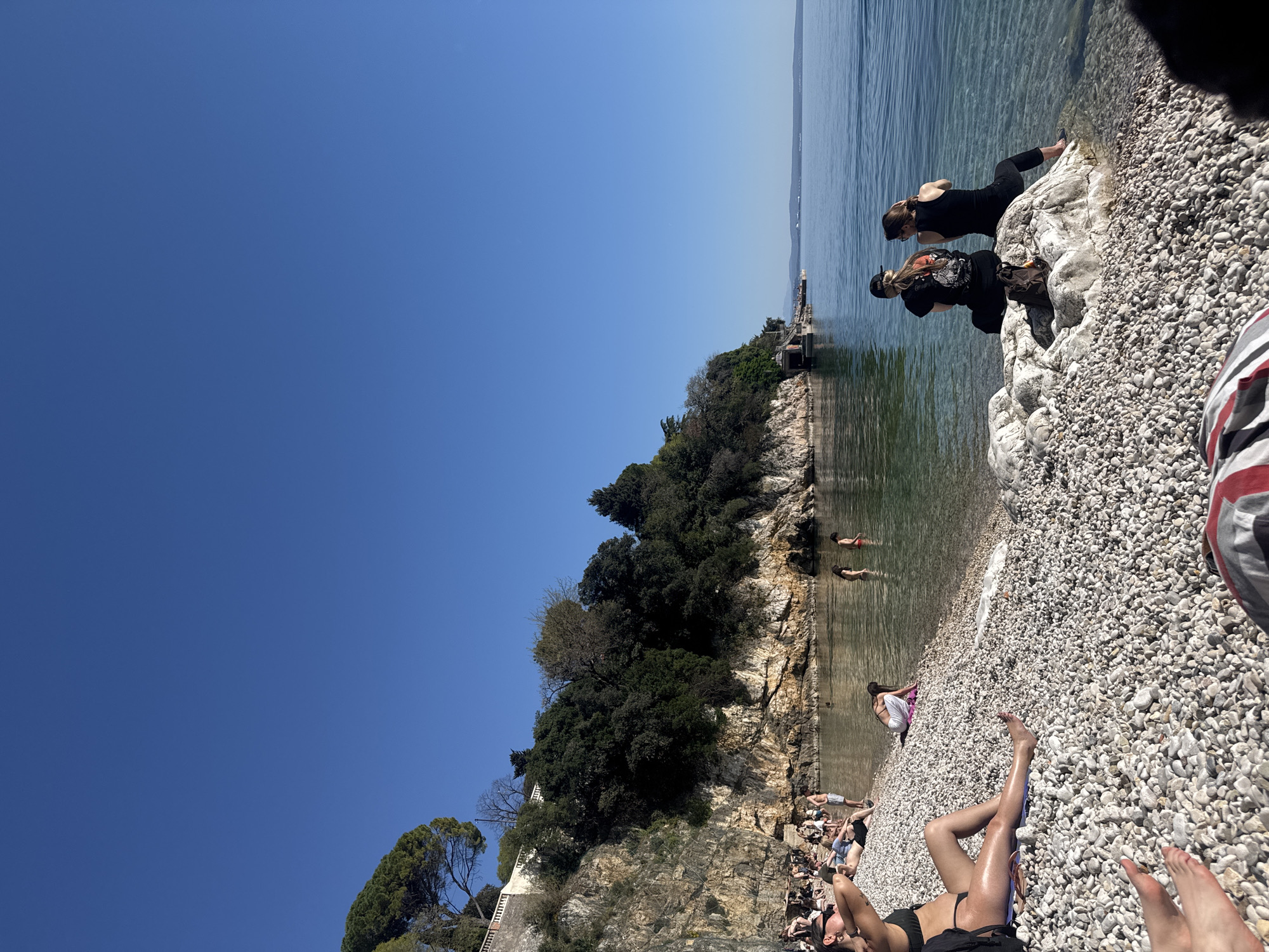 The image depicts a beach scene at Sablićevo Beach in the city of Grad Rijeka, Croatia, a pebble beach. A clear blue sky is overhead. Two women are sitting at the end of the beach next to a small pier. One woman is wearing black clothing and is sitting down. The other woman is standing. In the background, two other people are swimming in the water. On the beach itself, several people are sunbathing and relaxing. In the foreground, a woman is lying on the beach with her legs stretched out. The scene conveys a leisurely and sunny day at a coastal beach.