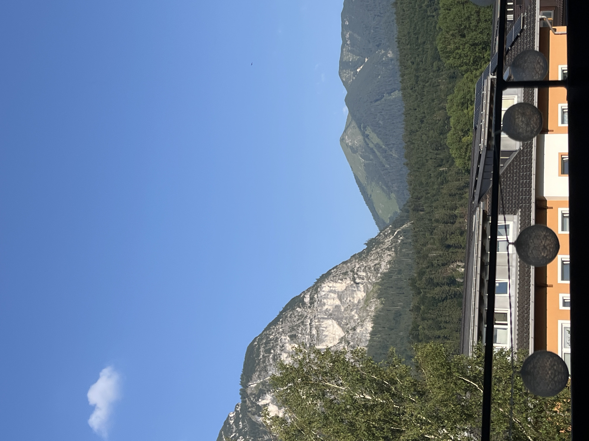 The image depicts a mountainous landscape with a clear blue sky. In the foreground, there are rooftops of buildings, some with solar panels. A tree with green foliage is visible on the left side. The midground features a dense forest that extends up the slopes of the mountains. The background is dominated by rugged, rocky mountains with patches of green vegetation. The overall scene suggests a rural or semi-rural setting, possibly on the outskirts of Garmisch-Partenkirchen, Germany, given its proximity to the Alps. There are no people or animals visible in the frame. The lighting indicates it is daytime, likely morning or afternoon due to the clear sky and shadows. The composition is framed from a slightly elevated perspective, looking out over the buildings towards the mountains.