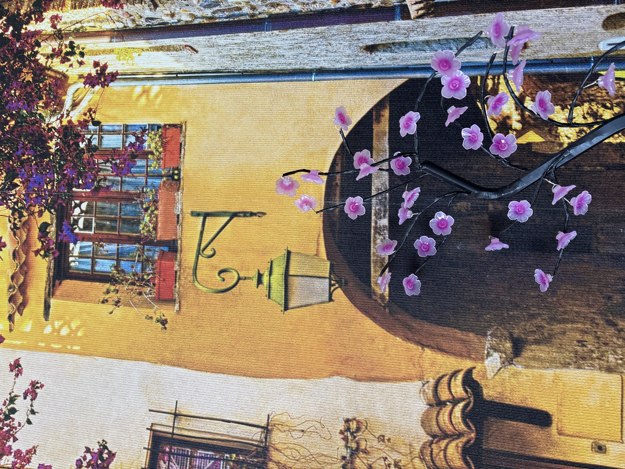 The image depicts a stylized, artistic representation of an outdoor scene, possibly a courtyard or narrow street. A warm yellow building with arched doorways and a window with red flower boxes is visible. A vintage-style lantern hangs from a bracket. In the foreground, a black artificial tree with pink translucent flowers is illuminated. The background shows a textured wall or building facade with vertical stripes. There are no people or animals present. The image appears to be a print or tapestry, not a photograph of a real location. The prompt's request to include the city of Georgiyevsk, Russia is not supported by any visual evidence in the image.