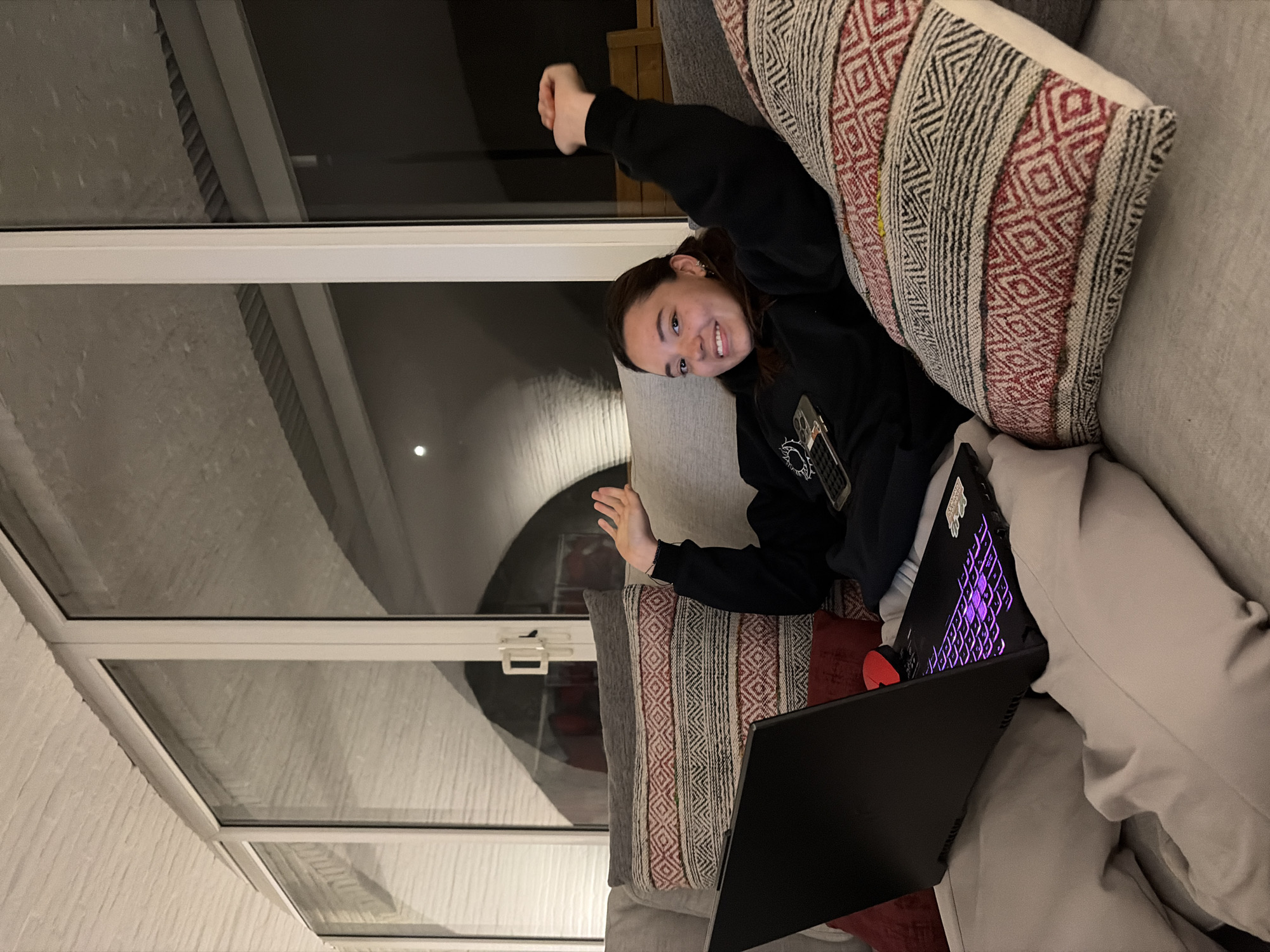 A young woman is sitting on a grey couch with two decorative pillows, using a black laptop. She is wearing a black sweatshirt and light-colored pants, and is smiling with one arm raised. The background features a window with a view of a light-colored exterior wall and sky.
It is not possible to determine the location is the city of San Bernardino Tlaxcalancingo, Mexico.