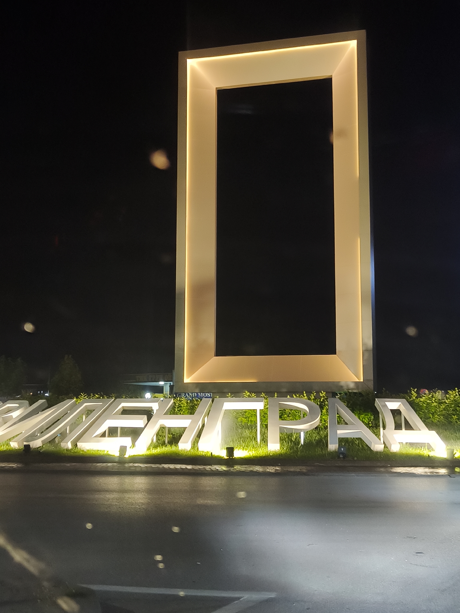 The media file captures an illuminated public monument or sign at night. The main subject is a tall, rectangular, open frame structure, brightly lit along its inner edges with warm yellow light. Below this structure, large white letters, also illuminated, spell out "СВИЛЕНГРАД" (Svilengrad in Cyrillic script). These letters are partially embedded in a bed of green foliage or small shrubs, which are also softly lit from below.
The scene is set outdoors at night, indicated by the dark sky. The ground in the foreground appears to be a paved road or asphalt surface, reflecting some of the light from the sign, suggesting it might be wet or highly polished. Several blurry circular lights, a bokeh effect, are visible across the dark background, possibly from distant streetlights or vehicle headlamps.
Towards the right, beneath the large frame and above the "ГРАД" portion of the text, a smaller line of text is faintly visible, reading "GRAND MOSI". The overall impression is a modern, well-lit welcome sign or landmark for the city of Svilengrad, Bulgaria, seen from a street-level perspective. No people are visible, and there is no specific activity taking place other than the display of the illuminated sign.