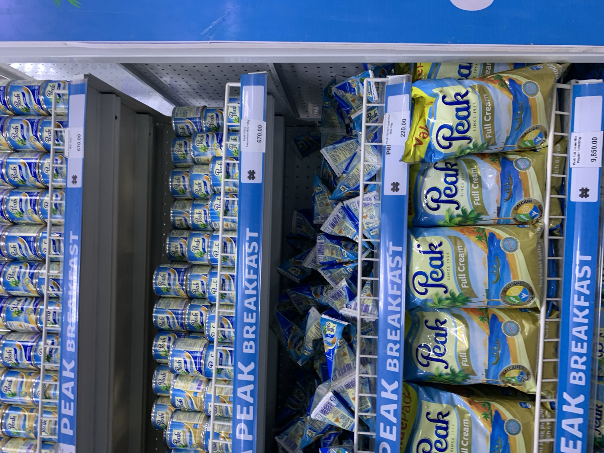 The image depicts a refrigerated display case in a retail store, likely in Ikorodu, Nigeria, showcasing Peak brand dairy products. The shelves are stocked with cans of Peak Filled Evaporated Milk (150g), priced at 670.00 (currency not specified). Next to the cans are pouches of Peak Filled Evaporated Milk (50g) at 220.00, and Peak Full Cream Milk Powder Refill (800g) at 9,850.00, all neatly arranged and facing forward for customers. The shelves are marked with blue branding strips reading "PEAK BREAKFAST."