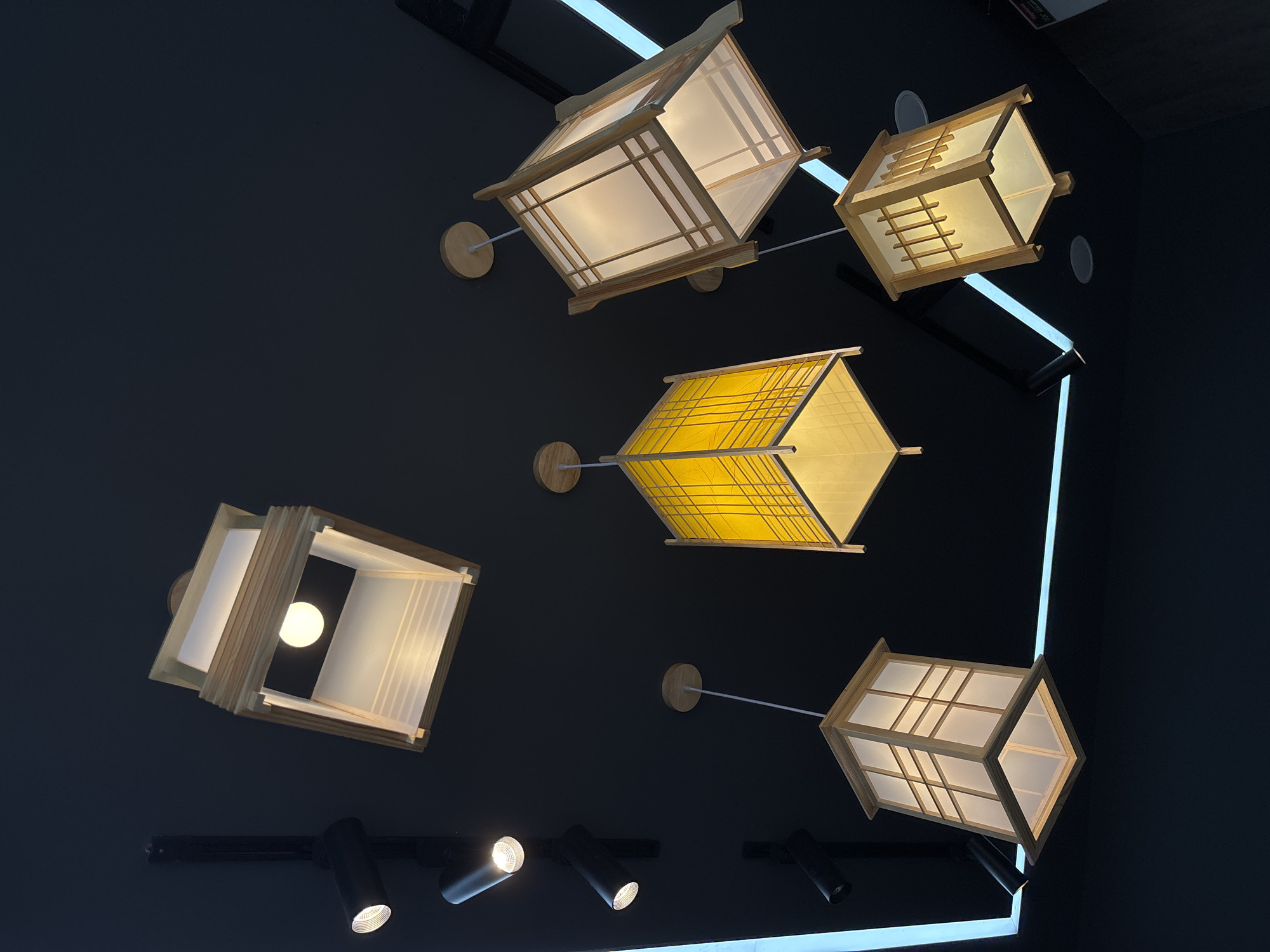 The image displays an interior view of a contemporary space, likely a ceiling, adorned with an assortment of lighting fixtures against a dark, possibly black or deep grey, background. The primary subjects are five geometric, lantern-style pendant lamps. Four of these lamps feature frames made of light-colored wood (appearing as natural wood or bamboo) with translucent white or cream-colored panels, arranged in a grid pattern. One distinctive lamp showcases bright yellow translucent panels, also with a fine grid pattern. These lamps are suspended at different heights and angles by slender rods attached to circular wooden mounts on the ceiling.
Below and around these hanging lanterns, black cylindrical track lights are visible, mounted on a track system, with some actively emitting light. Additionally, bright, linear LED light strips are integrated into the architecture, contributing to the overall illumination and outlining parts of the space.
The scene focuses exclusively on the lighting design; no people or discernible activities are present. The combination of traditional Asian-inspired lantern designs with modern track lighting and minimalist linear lights creates a sophisticated and warm ambiance, suggestive of a commercial establishment, hotel lobby, or upscale residence. The lighting indicates an indoor setting where artificial light is the primary source, likely during the evening. The design elements are consistent with contemporary aesthetics found in places like Đà Nẵng, Vietnam. No legible text is visible in the image.