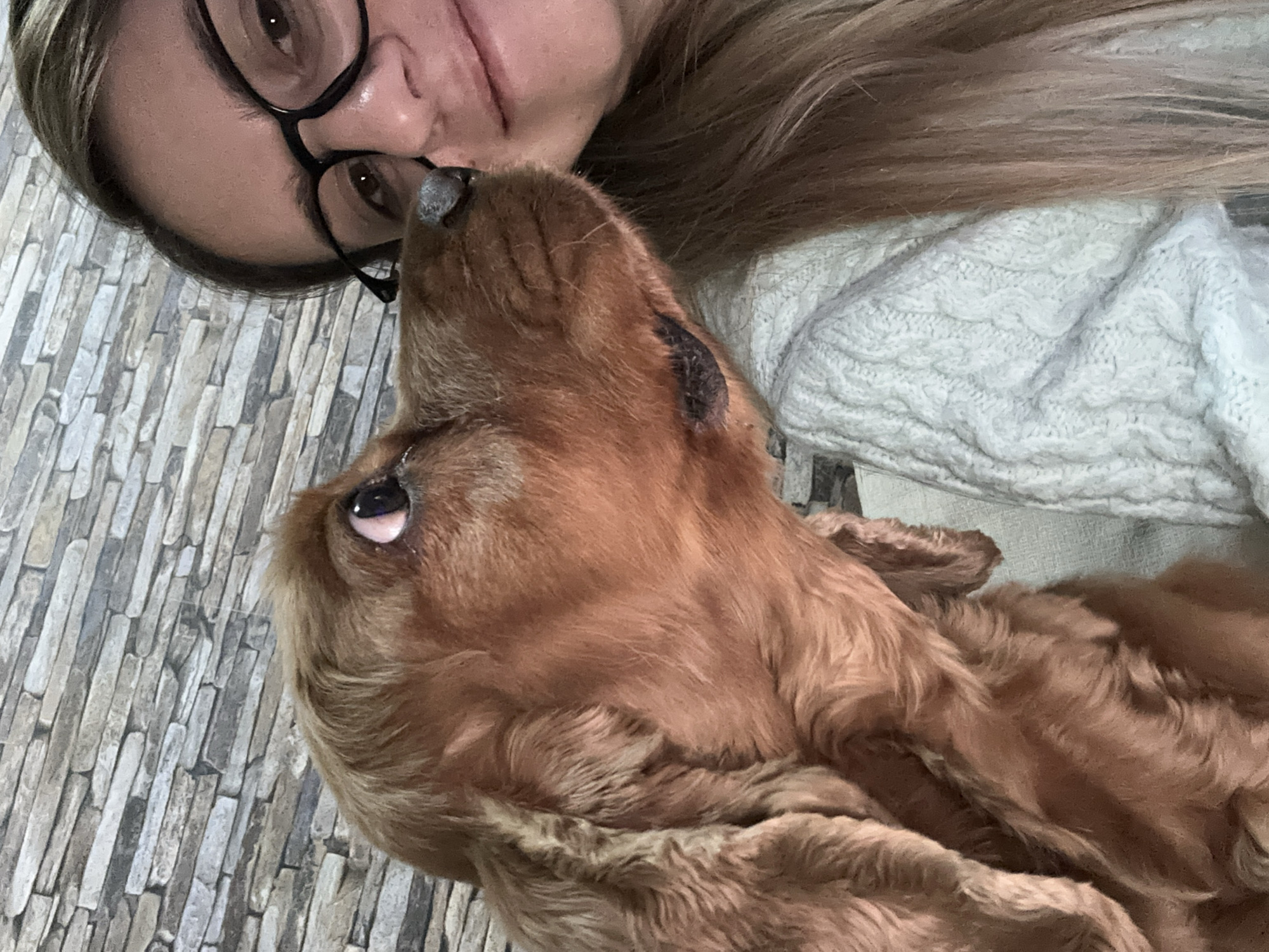 A woman with glasses and light hair is lying down, her face close to a reddish-brown Cocker Spaniel dog. The dog is looking up towards the woman's face. The woman is wearing a light-colored cable knit sweater. The background appears to be a wall with a stone tile pattern. There is no information in the image to suggest it is taken in Sofia, Bulgaria.