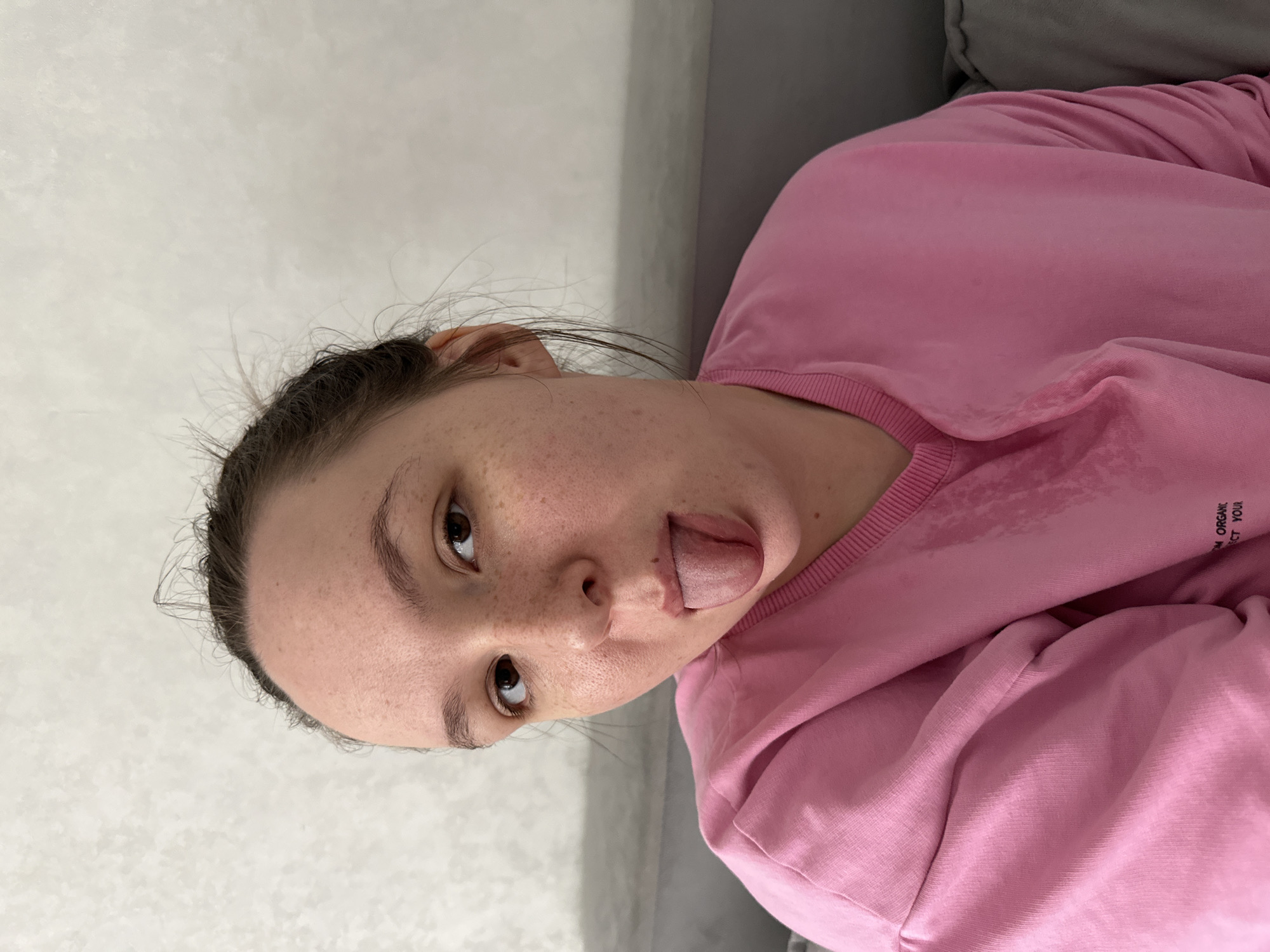 A young woman with brown hair pulled back and light skin with freckles is shown from the chest up, looking at the camera with her tongue sticking out. She is wearing a pink sweatshirt, which appears damp in places on the front. The background is a plain, textured wall. There are no other people or objects visible in the image. The image appears to have been taken indoors. The location is not specified in the image.