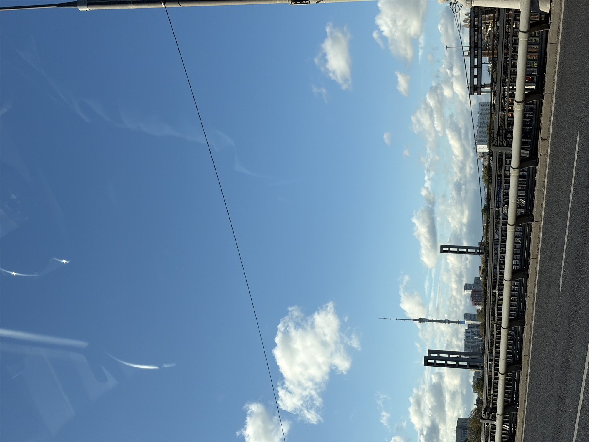 A view of the Ostankino Tower in Moscow, Russia, is visible in the distance under a clear blue sky dotted with white clouds. The tower is a prominent landmark, appearing as a tall, slender structure with a pointed spire. In the foreground, a bridge railing with a metal fence runs horizontally across the image, separating a road from the cityscape beyond. The road itself, made of asphalt, is visible in the lower part of the frame with white lane markings. Other structures, including buildings and what appear to be industrial elements, can be seen along the horizon line, partially obscured by the bridge and vegetation. The overall scene suggests a day view from a moving vehicle, possibly on a bridge, looking towards the iconic tower and the city skyline.