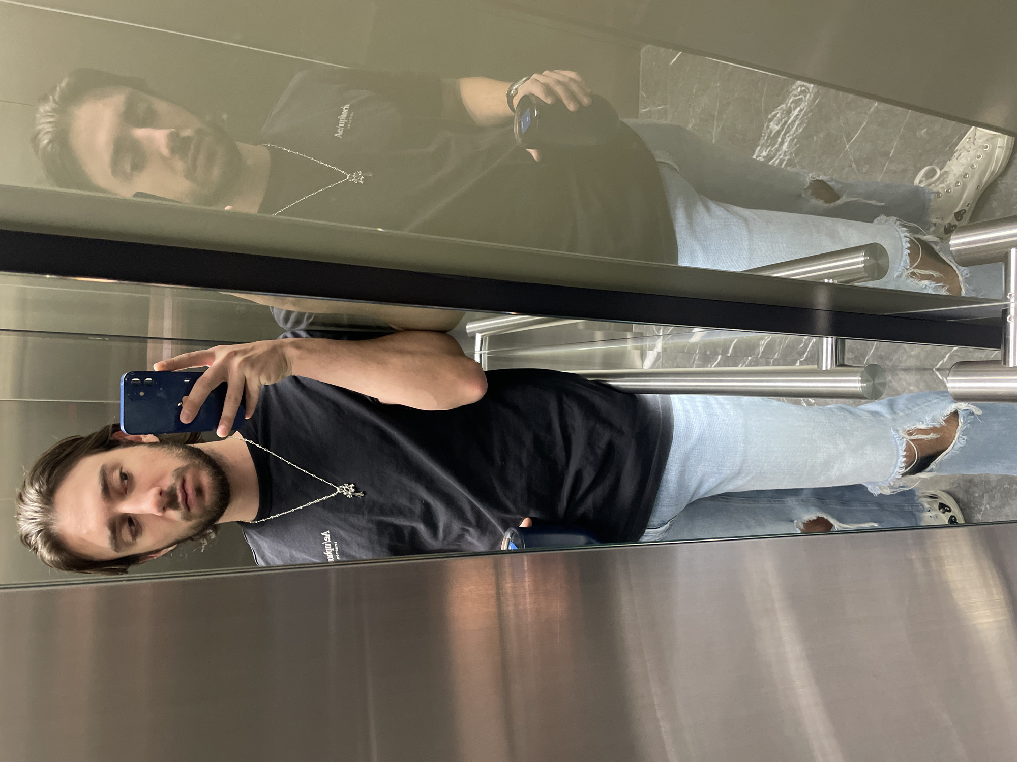A fair-skinned male with dark hair and a beard is taking a selfie in what appears to be an elevator. He is wearing a black t-shirt with white lettering on the front, ripped light blue jeans, and white sneakers. A silver necklace with a pendant hangs around his neck. He is holding a blue smartphone in his left hand to take the picture. The elevator has stainless steel walls and a grab bar. The image also shows a reflection of the man in the elevator wall. The specific location within Mexico City is not discernible from the image.