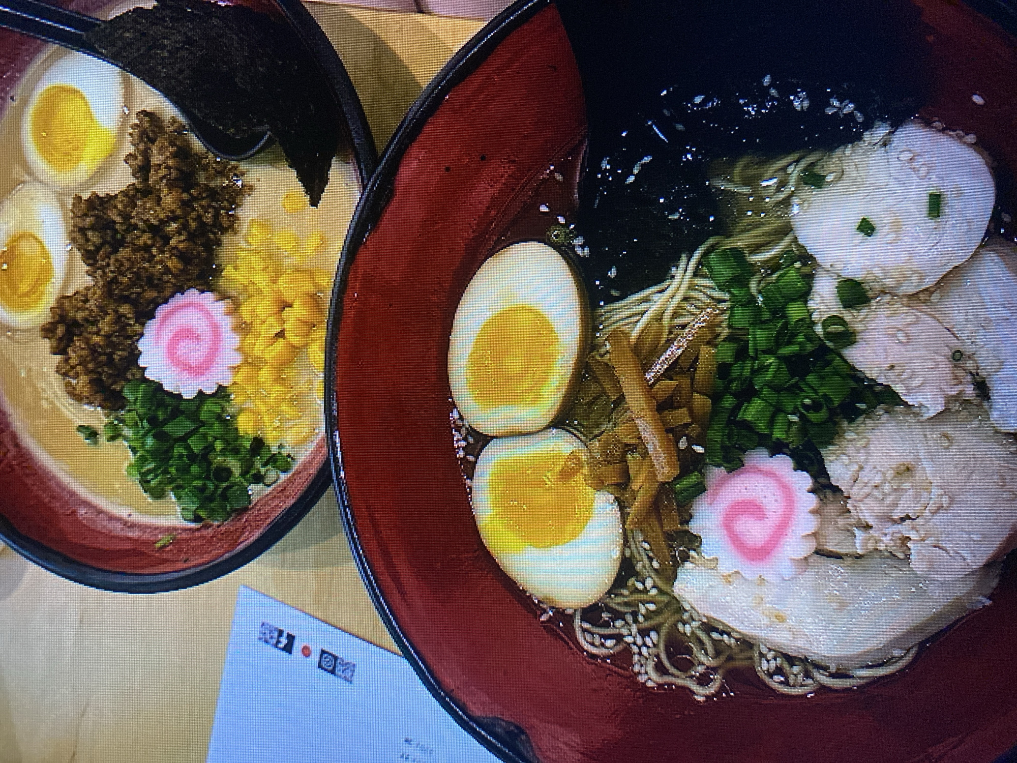 The image shows two bowls of ramen noodles. The bowls are red on the inside with black rims. The ramen in the bowls contains broth, noodles, sliced hard-boiled eggs with yellow yolks, green onions, corn, a pink and white swirled fish cake, and what appears to be seasoned ground meat and nori in the upper bowl. The lower bowl also contains sliced chicken and bamboo shoots. There is a piece of paper with Japanese characters and a red dot, likely a menu, visible to the left of the bowls. The setting appears to be a restaurant. Based on the visible text, the restaurant is likely "YO! Sushi" located at Plac Wolności 9, 61-713 Poznań, Poland.