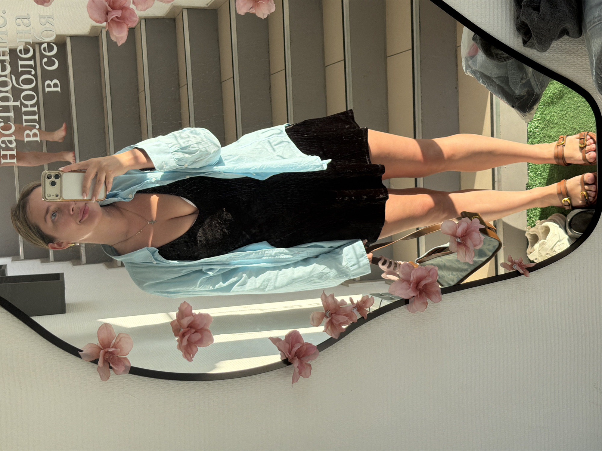 A young woman is captured in a mirror selfie, seen from a perspective that has the image rotated 90 degrees clockwise. She smiles subtly at the camera of her light-colored smartphone, which she holds up. Her hair is pulled back, and she wears a black mini-dress, a light blue unbuttoned long-sleeved shirt over it, and brown sandals with gold buckles. A delicate cross pendant necklace adorns her neck.
The mirror has an irregular black frame decorated with several pink artificial flowers. Reflected in the mirror's background are what appear to be light-colored stairs or steps, possibly made of concrete or tile, suggesting an outdoor area or a multi-level interior. A patch of green artificial turf is visible in the upper right of the reflection, along with some folded dark clothes or items. Bare legs and feet are also faintly reflected, possibly from another person or a complex reflection.
Prominently displayed on the mirror's surface is Cyrillic text that reads: "Настроение влюблена в себя," which translates to "Mood in love with myself." The scene is brightly lit, indicating it is daytime. The overall impression is one of a self-appreciative moment in Chaikovsky, Russia.