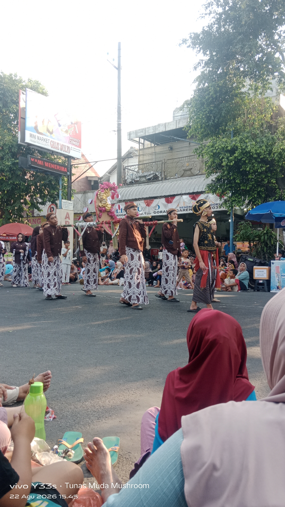 In Genengan, Indonesia, a street procession is taking place, captured from a low angle. The foreground shows the bare feet and legs of several spectators sitting on the asphalt, with a green water bottle and flip-flops nearby. The middle ground is occupied by a group of young men in traditional Indonesian attire, carrying a decorated palanquin on their shoulders. They are walking in a line, with one individual in more elaborate headwear and a dark, gold-embroidered vest leading the way. Behind them, other participants and spectators are visible, with some seated on the side of the road. The background features buildings with signage, including a "Mini Market" billboard, and trees. The scene appears to be a cultural event or festival, with the procession moving down a street lined with onlookers.
