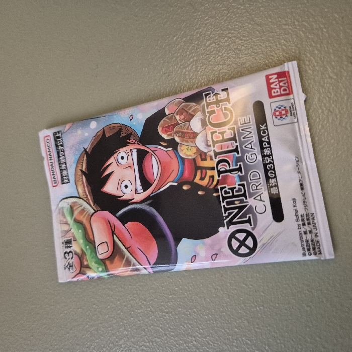 A sealed foil pack of the "One Piece Card Game," specifically the "Strongest 3 Brothers Pack," rests on a flat, light grey-greenish surface. The pack's front features a vibrant illustration of Monkey D. Luffy, wide-mouthed and smiling, wearing a straw hat and black jacket, holding a large piece of meat. Additional food items are depicted to his left. Text on the pack includes "BANDAI NAMCO," "ONE PIECE CARD GAME," "MADE IN JAPAN," and copyright information for Eiichiro Oda. The surface displays a subtle, granular texture with even illumination. There are no individuals visible, and no actions or interactions are occurring. The image provides no identifiable contextual information regarding its geographical location within Strasbourg, France, or any other specific city.