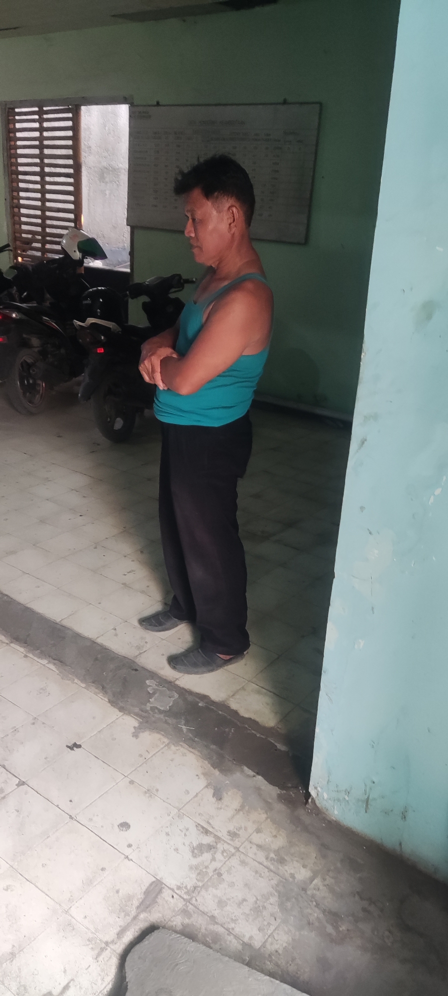 A middle-aged man of Asian descent stands with his arms crossed, facing to the left of the frame. He is wearing a teal tank top and black pants. He is standing on a tiled floor that appears wet and has some dirt markings. To his left, there are two dark-colored motorcycles. Behind the motorcycles is a window with horizontal wooden blinds. A whiteboard with writing is visible on the wall behind the man. The environment appears to be an indoor or semi-covered area, possibly a garage or storage space, with a light green wall to the right of the man. The scene is set in the city of Gebang, Indonesia.