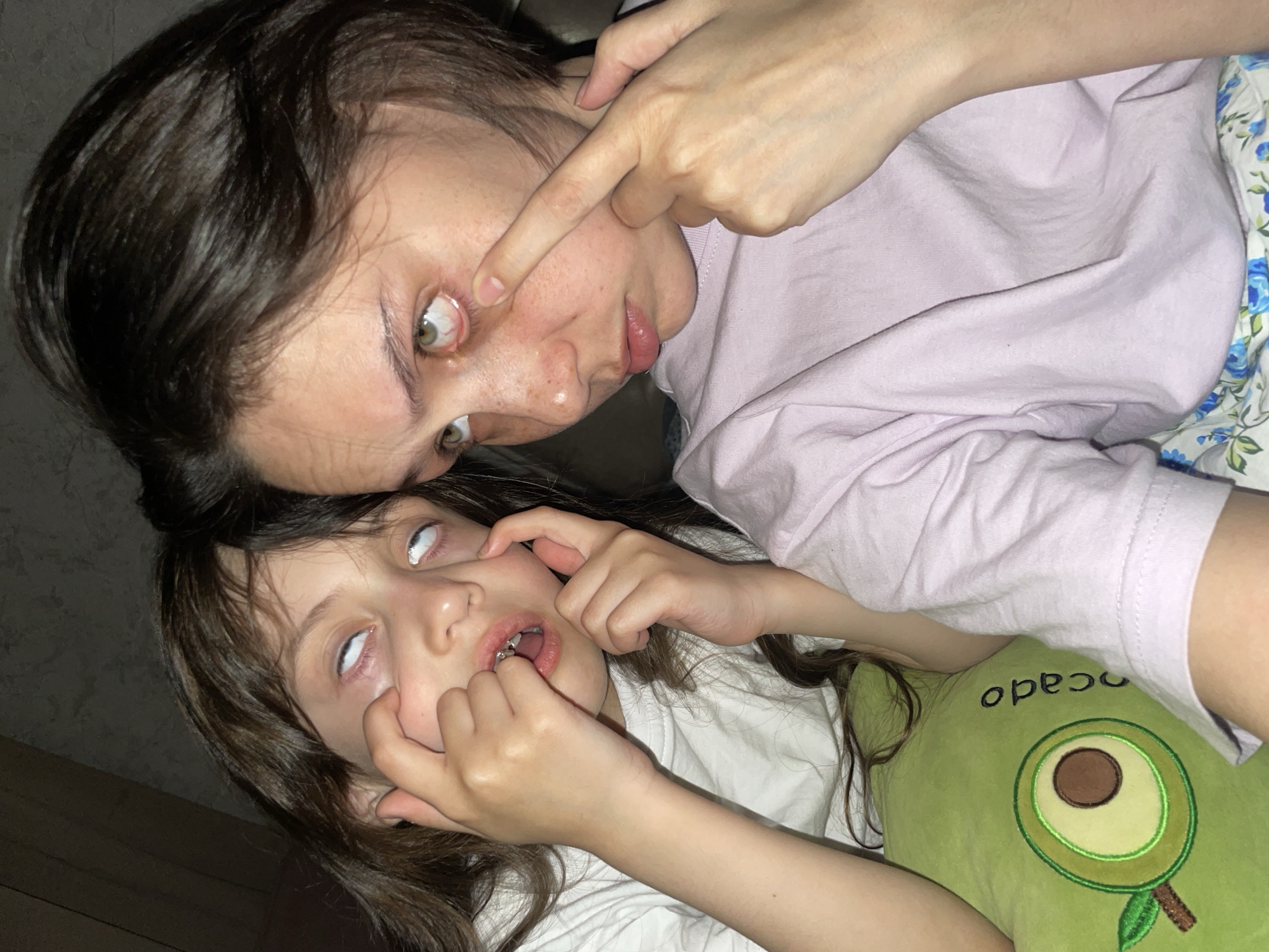 Two individuals are visible in the image, seemingly posing for the camera. The older individual is partially angled, facing the camera with one hand near their eye. They have dark hair and are wearing a light-colored top. The younger individual is lying on a pillow shaped like an avocado. The child has dark hair and is wearing a white shirt. The pupils of the child are rolled back. The background appears to be indoors. The image does not contain any information that could be used to identify the location as Izhevsk, Russia.