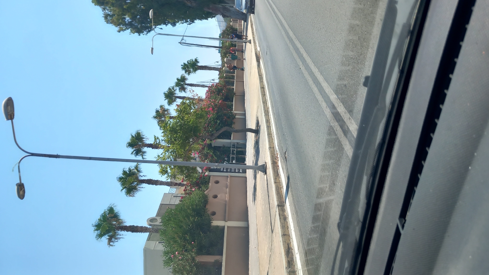 The image shows a street scene in Kos, Greece, on a clear, sunny day. In the foreground, a car's dashboard and windshield wipers are visible, suggesting the photo was taken from inside a vehicle. The road ahead has double white lines and a paved sidewalk. Palm trees and lush green bushes line the sidewalk, interspersed with flowering plants. A beige-colored wall with circular accents runs along the property line. Several people are visible walking and riding scooters on the sidewalk. In the background, a building with a modern architectural style can be seen. Streetlights with dual fixtures are present along the sidewalk. A dark-colored vehicle is parked on the right side of the road.