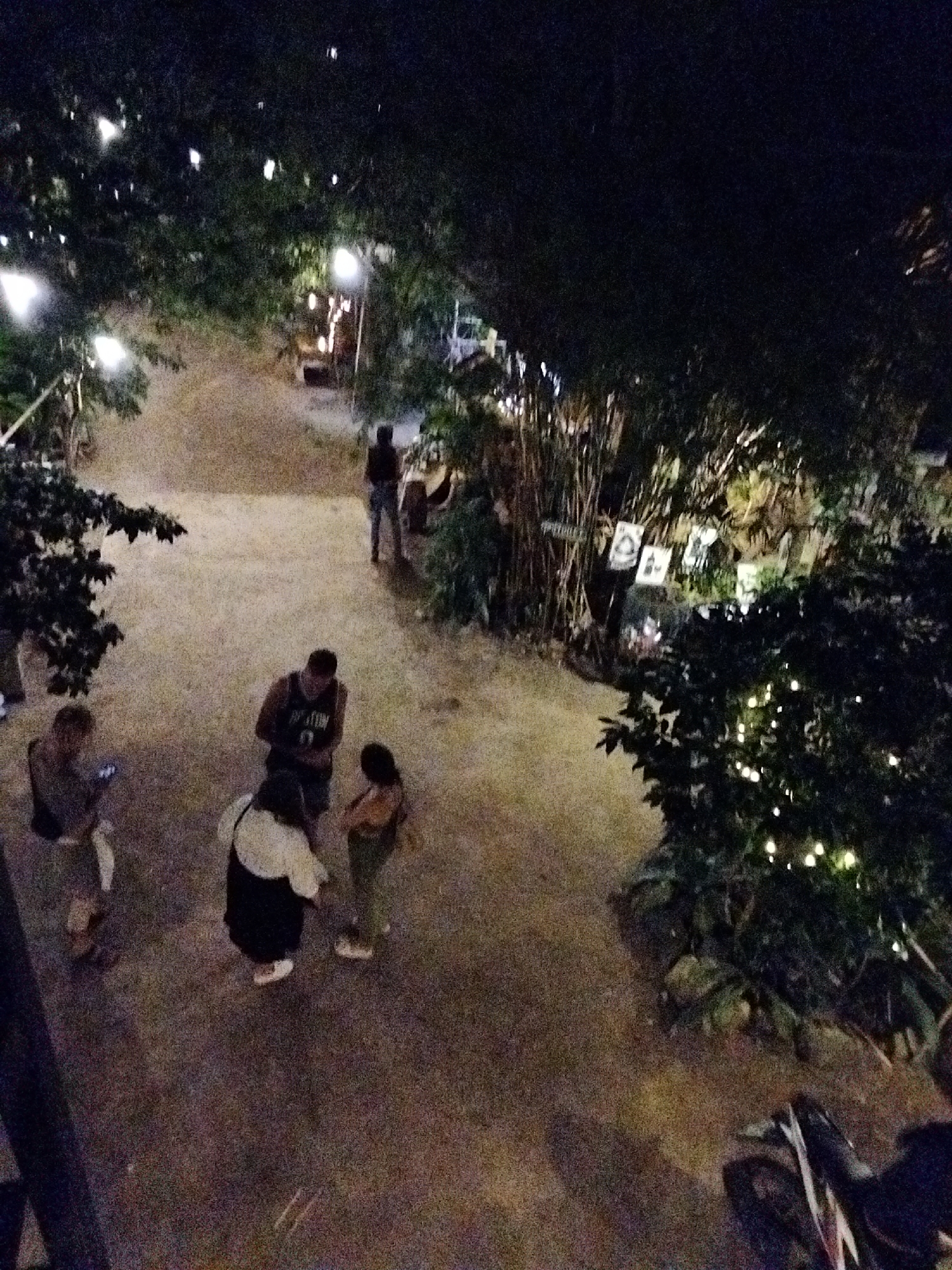 The image shows an outdoor scene at night in ตำบลเกาะพะงัน, Thailand. Several people are visible, including a group of three standing close together, and another person walking away in the background. The area is lit by string lights hanging in trees, as well as lanterns. Some pictures are hung on the bamboo behind the group of people standing together. The ground is dirt or sand.