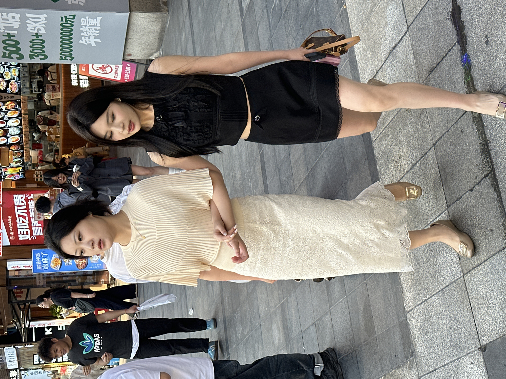 This image captures a street scene in an urban commercial area, likely in Liangjiang, China. The primary subjects are two young women standing on a paved sidewalk, facing generally forward and slightly rightward from the viewer's perspective.
The woman positioned closer to the viewer is dressed in a sleeveless black top with a textured or embroidered bust area, paired with a short black skirt featuring a delicate, scalloped lace hem. She has long, dark hair parted in the middle and is holding a small, patterned handbag and a mobile phone in her right hand. She wears light-colored flat shoes with a gold buckle. Her head is tilted downwards, and her expression appears calm.
The woman slightly behind and to her left is wearing a light beige, vertically ribbed top and a long, light beige lace skirt. Her dark hair is styled in a bob, and her hands are clasped gently in front of her. She also wears light-colored flat shoes with a gold buckle, similar to her companion. Her expression is neutral and somewhat pensive.
In the background, other people are visible walking and standing, suggesting a bustling environment. The setting features various commercial establishments, including restaurants and shops, with brightly lit displays of food visible in some windows. The time of day appears to be daylight, with clear weather.
Visible text on signs further clarifies the commercial nature of the area. One prominent sign displays "500+店" (500+ stores), "3000+SKU", "500000000元", and "年销量" (annual sales), indicating a large-scale business or shopping complex. Other signs feature Chinese phrases such as "好吃不贵" (delicious and inexpensive), "解暑神器" (summer heat relief artifact), "营养美味" (nutritious and delicious), "冰麻了" (iced and numbing/frozen), "禁止摩托车通" (no motorcycles allowed), and operational hours "9.00-23.00". A man in the background is seen wearing a black t-shirt with an "ADIDAS" logo.