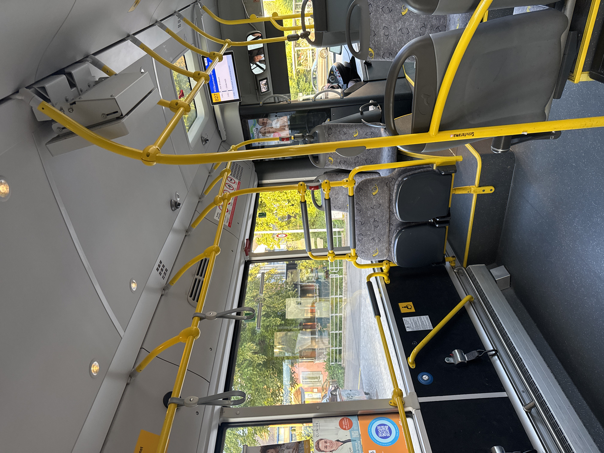 The image captures the clean and modern interior of a public bus, likely in Fehraltorf, Switzerland, during a sunny daytime. The view is from the rear of the vehicle, looking forward towards the driver's area and the front exit.
**Subjects and Objects:** The bus interior features bright yellow grab bars and poles for passengers, crisscrossing the ceiling and lining the aisle. Grey and black seating is arranged along both sides, with some seats appearing to be fixed and others potentially foldable, one bearing the text "Scouterama." The floor is dark, with patterned sections. Large windows offer views of green trees and distant buildings, indicating an outdoor, possibly suburban, environment. The ceiling has several recessed lights and ventilation grilles. Towards the front, a digital display screen is visible, along with smaller screens or mirrors and a partial view of the driver's seat. A dedicated space, likely for wheelchairs or prams, is equipped with a safety belt, a blue button, and a yellow support bar, accompanied by a wheelchair symbol. An advertisement on one window shows a person's image, a QR code, and text.
**Activity or Event:** The bus appears to be empty of passengers, suggesting it might be at a stop, between routes, or awaiting service. No specific activity by individuals is depicted.
**Notable Details:** The bright sunlight filtering through the windows indicates it is daytime with clear weather. The modern design and clean state of the interior suggest a well-maintained public transport vehicle. The presence of German text ("Willkommen" on the display and "von Zürich" on an advertisement) aligns with the provided location context of Fehraltorf, Switzerland.
**Visible Text:** Key visible text includes "827 Willkommen/W" on the overhead digital display, "Scouterama" on a seat, and various fragments from an advertisement such as "CLINIUS," "50 Min von Zürich," "re erste suchung," and "net."