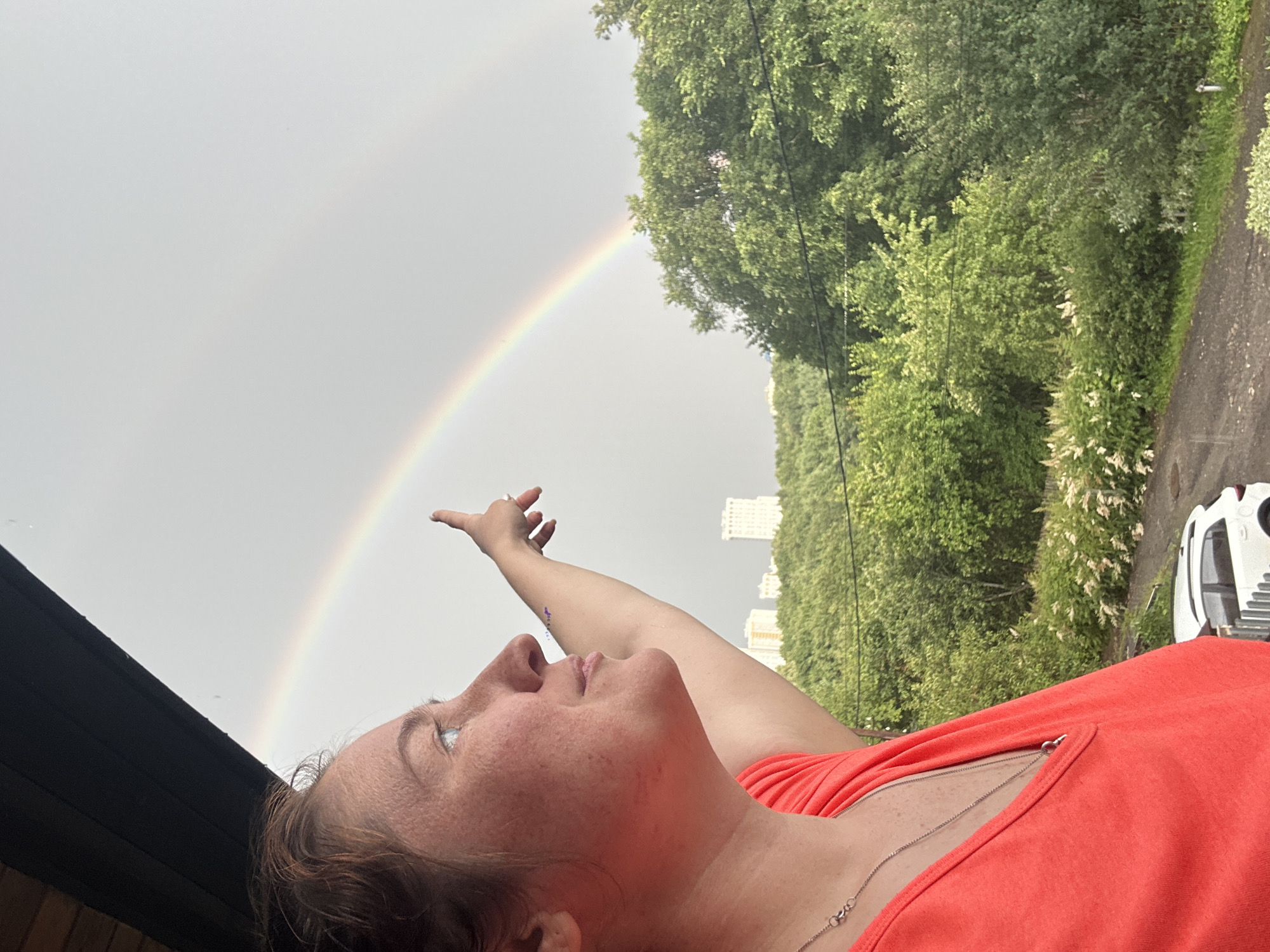 In Nizhny Novgorod, Russia, a woman with reddish-brown hair is captured in a side profile, her face illuminated by natural light. She is wearing an orange t-shirt and a silver necklace, and is looking upwards and to her right. Her right arm is raised, and her index finger is extended as if pointing at something in the sky.
The sky is overcast, with a faint rainbow visible, arcing across the gray expanse. The upper portion of the image is framed by a dark overhang, possibly a balcony or roof.
Below the sky, a lush green landscape unfolds. Dense trees with vibrant green foliage dominate the mid-ground. In the distance, several tall, light-colored buildings rise above the trees, suggesting an urban or suburban setting. The foreground shows a paved area, a white car, and some white flowering bushes. The overall scene evokes a moment of observation of a natural phenomenon within a city.