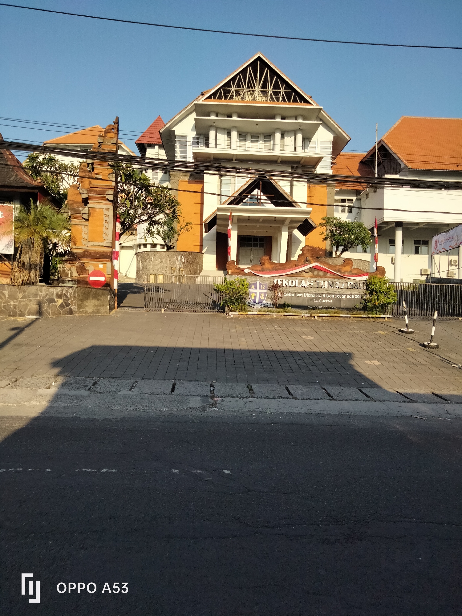 The image shows the entrance to a school building in Denpasar, Indonesia. The building has an orange and white facade with a distinctive multi-level roof structure. In front of the building, there is a sign that reads "SEKOLAH TUNA DAU" in large letters, with additional text below indicating the address and contact information. The school's grounds are paved with interlocking bricks, and a metal fence encloses the property. To the left of the entrance, there is a traditional Balinese stone gate, a palm tree, and a red and white striped pole with a red circular sign indicating "no entry." Several power lines are visible against the clear blue sky. The sun is casting long shadows on the paved area, indicating it is likely morning or late afternoon. The scene appears quiet, with no people visible in the frame.