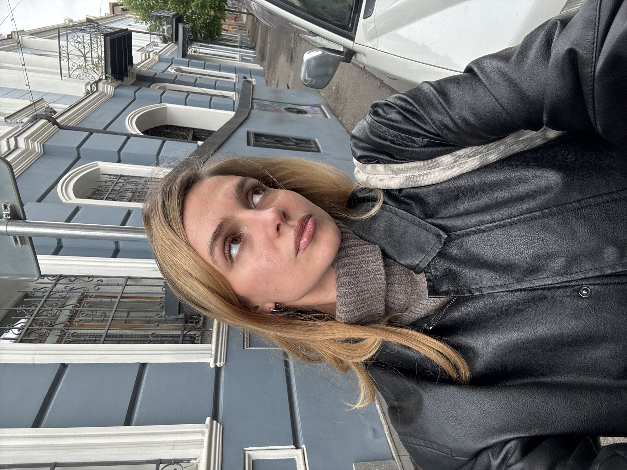 A woman with blonde hair, wearing a black leather jacket over a neutral-colored sweater, poses outdoors on a street in Tbilisi, Georgia. The architecture in the background is characterized by blue and white buildings with ornate window frames and ironwork balconies. A white vehicle is partially visible on the right side of the frame. The woman is looking upward and to the left, and the photo is taken at an angle.