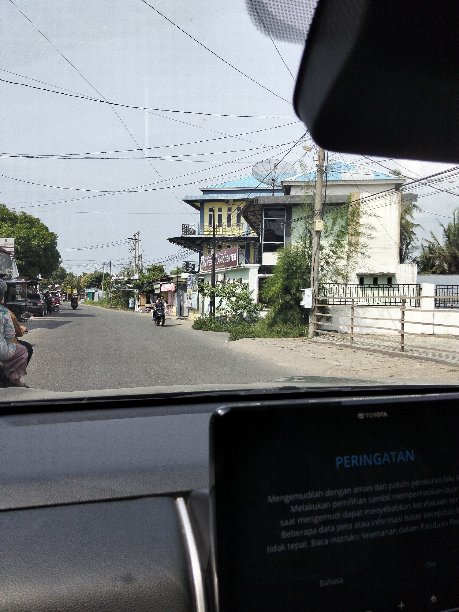 The image depicts a street scene in Geulanggang Teungoh, Indonesia, viewed from the interior of a vehicle. The foreground shows the dashboard of a car with a screen displaying a warning message in Indonesian. The screen reads "PERINGATAN" (Warning) and provides instructions for safe driving, mentioning "Mengemudilah dengan aman dan patuhi peraturan lalu li" (Drive safely and obey traffic regulations), "Melakukan pemilihan sambil memperhatikan layar li saat mengemudi dapat menyebabkan kecelakaan ser" (Selecting while looking at the screen while driving can cause serious accidents), "Beberapa data peta atau informasi batas kecepatan ter tidak tepat. Baca instruksi keamanan dalam Panduan Per" (Some map data or speed limit information may not be accurate. Read the safety instructions in the User Manual), and "Oke" (OK) and "Bahasa" (Language) at the bottom.
The middle ground shows a paved road with several motorcycles and a car. Two people are visible on one of the motorcycles, and another person is seated on a motorcycle behind them. Further down the road, there are buildings on both sides. On the right, a multi-story building with a light green roof and white walls is prominent. A fence with wooden posts and metal bars runs along the front of this building. On the left side of the road, there are smaller shops and trees. A sign on one of the buildings reads "ZAWIEH SLAMIC CENTER." Overhead, numerous power lines crisscross the sky.
The background consists of a cloudy sky and more distant buildings and trees. The overall impression is of a typical street in a populated area with ongoing activity.