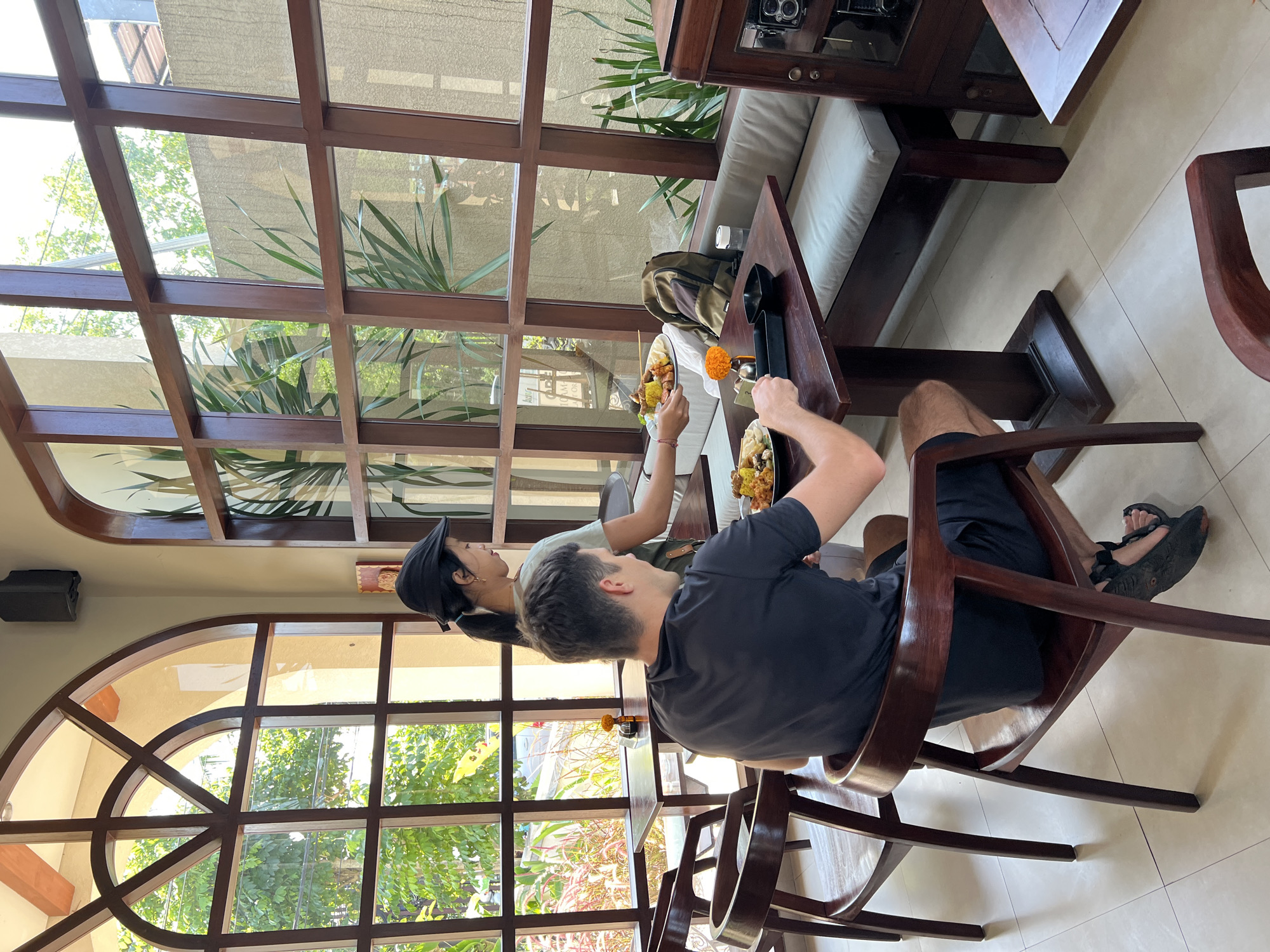 In Ubud District, Indonesia, a woman in a black cap and a light-colored shirt, possibly a server, is placing a plate of food on a dark wooden table. A man in a black t-shirt and dark shorts is seated in a dark wooden chair, facing away from the camera. Another dark wooden chair is visible in the foreground. The scene is set indoors, with large windows showcasing lush greenery outside. The restaurant appears to have a rustic ambiance with its wooden furniture and decor. The table is set with plates of food, a black bowl, a small orange flower, and a bottle of water. A backpack rests on a bench to the right of the table.