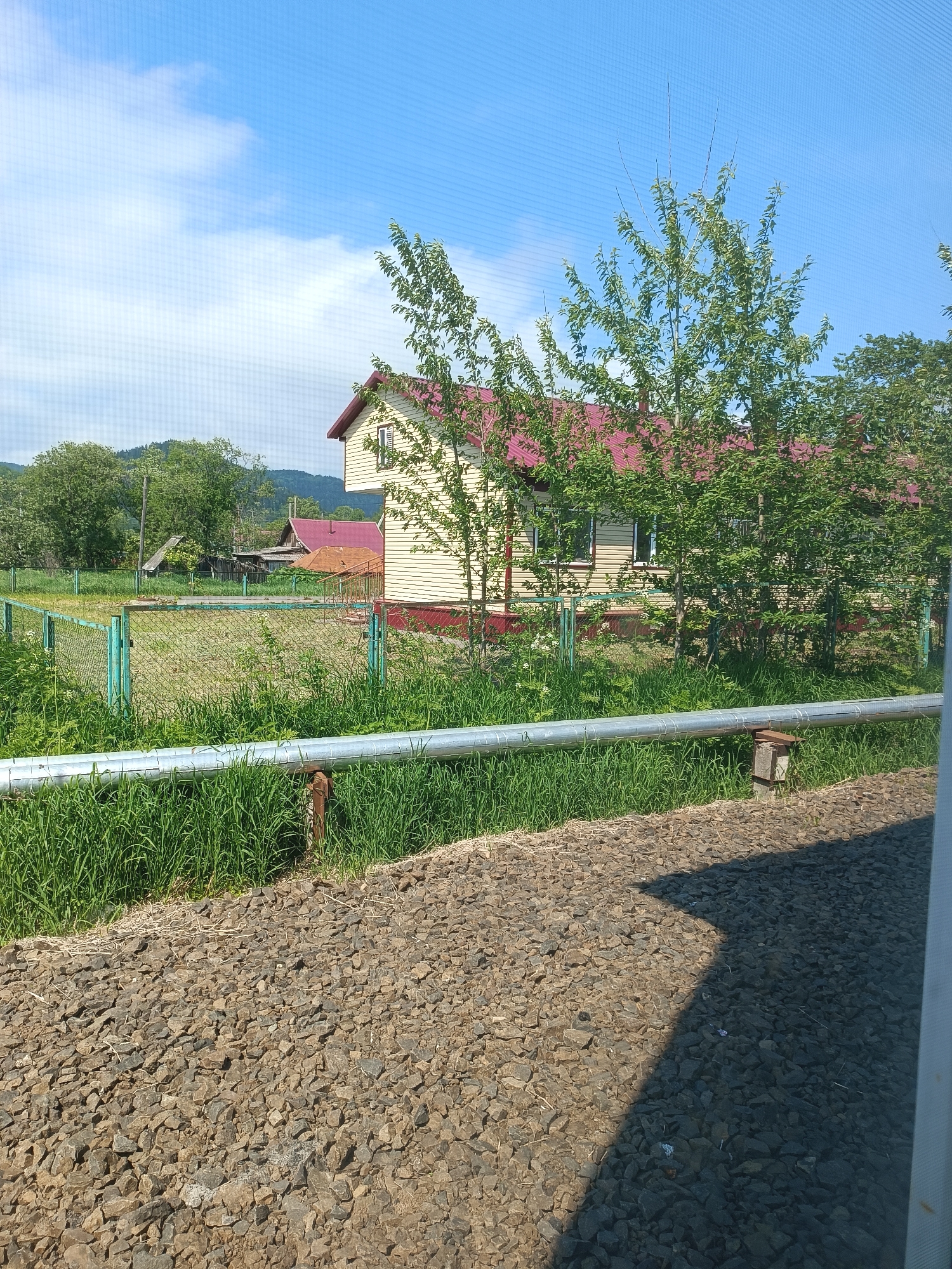 The image shows a rural scene in Krasnopol’ye, Russia, on a sunny day. In the midground, a light-colored house with a red roof stands behind a green chain-link fence. The house has visible windows and appears to be a dwelling. Adjacent to the house are trees with green foliage. In the foreground, a metal pipe runs horizontally across the frame, supported by rusty posts. The ground in the foreground is covered with dark-colored gravel. Behind the fence, there is a patch of green grass and other vegetation, along with some outbuildings and a larger forested area with hills in the background. The sky is blue with some white clouds. The image appears to have been taken from an elevated position, possibly through a window or screen.