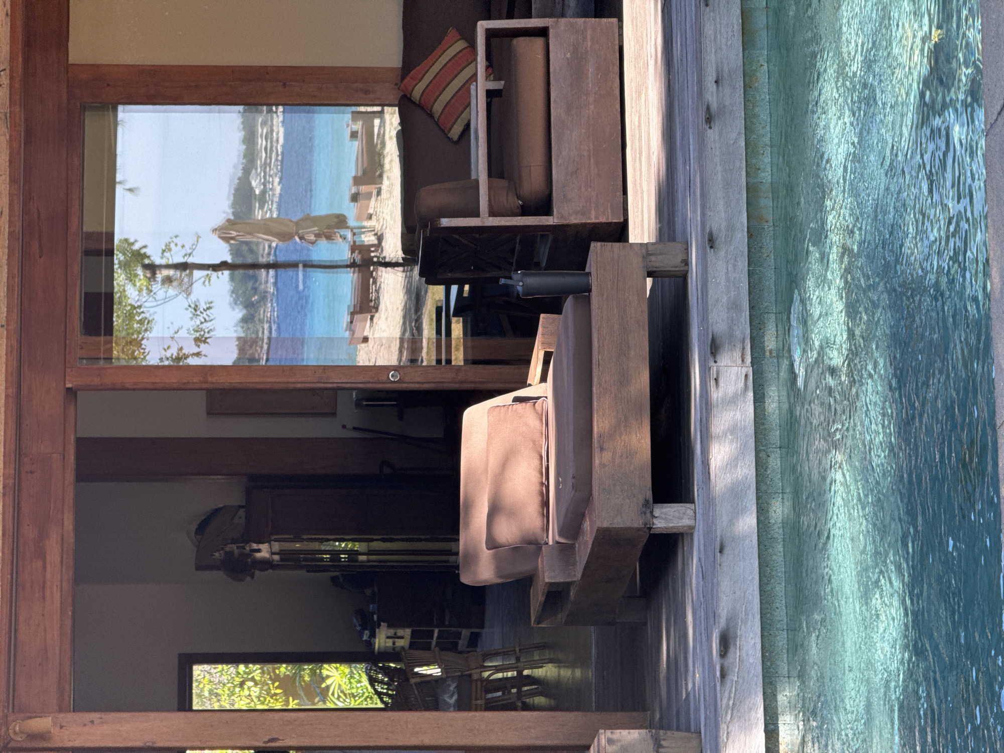 The image, when rotated upright, depicts a tranquil resort scene, likely a private villa or bungalow, combining indoor comfort with outdoor natural beauty. In the foreground, a light grey wooden deck runs alongside a clear, turquoise swimming pool or water feature, its surface gently rippled by sunlight. Visible on the deck are sturdy, dark wooden lounge chairs or daybeds with brown cushions, one of which has a dark cylindrical attachment.
Behind the deck, large glass sliding doors or windows separate the exterior from a softly lit interior space. Inside, visible through the glass, is an inviting seating area featuring wooden furniture, including a sofa with a distinctive red, orange, and beige striped cushion. Reflections on the glass offer a distorted but clear view of the outside.
Beyond the immediate outdoor area, a vibrant tropical landscape unfolds: a bright, sandy beach meets a calm expanse of blue and turquoise ocean, with distant land or hills under a clear sky. Faint outlines of beach umbrellas or loungers can be seen on the sand. Green foliage, possibly from trees or plants, is also reflected or visible through the lower sections of the glass.
The bright sunlight illuminating the water and deck, contrasting with the soft shadows cast by the furniture, suggests a sunny daytime setting. No individuals are present, and no specific activity is taking place, contributing to an overall atmosphere of peace and relaxation. There is no visible text in the image.