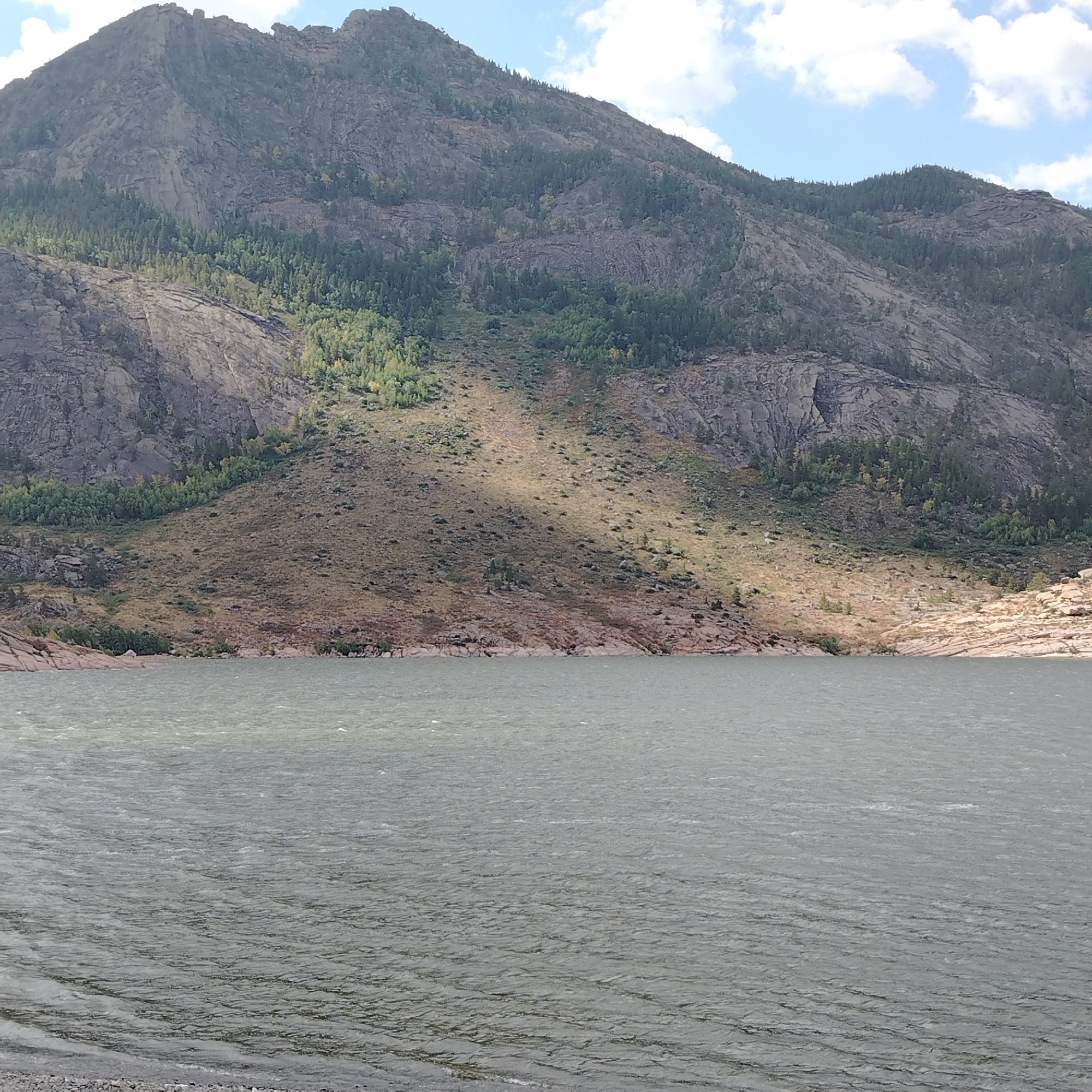 The image depicts a body of water in the foreground, with a large, rocky mountain rising behind it. The mountain is covered in sparse vegetation, with patches of trees and dry grass. The sky above is partly cloudy. The water is choppy, suggesting a windy day. The shoreline consists of rocks and pebbles. The scene appears to be a natural landscape. The location is in the vicinity of Toraygyrov, Kazakhstan, a region known for its mountainous terrain and lakes. There are no people or animals visible in the image.