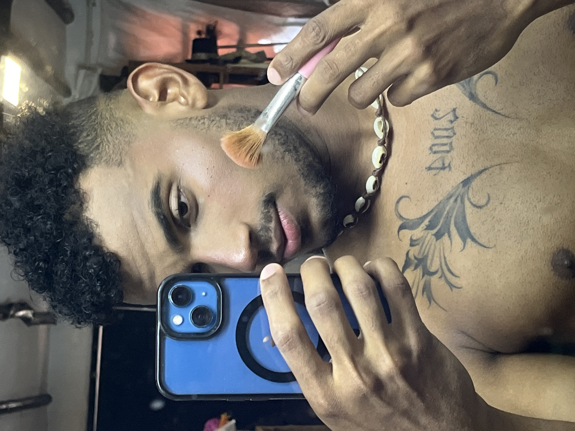 This detailed image, captured in Porlamar, Venezuela, depicts a young adult male taking a selfie in what appears to be an indoor setting. He is positioned in front of a reflective surface, likely a mirror, and is shirtless, revealing his bare chest. He has short, dark, curly hair with faded sides, and a full beard. His left hand holds a blue iPhone with a clear case and a visible black MagSafe ring, positioned to capture his reflection. The phone's dual rear cameras are distinctly visible. With his right hand, he holds a small makeup brush with a pink handle and light brown bristles, which is positioned against his right cheek, suggesting the application of makeup or highlighter. A necklace made of light-colored cowrie shells strung on a dark cord is worn around his neck. On his chest, two tattoos are visible: an ornate, dark-ink floral or leafy design on his left side, and the word "DOOS" rendered in an Old English font centrally on his upper chest. The background is a blurred reflection showing indistinct shapes and a bright light fixture at the top right.