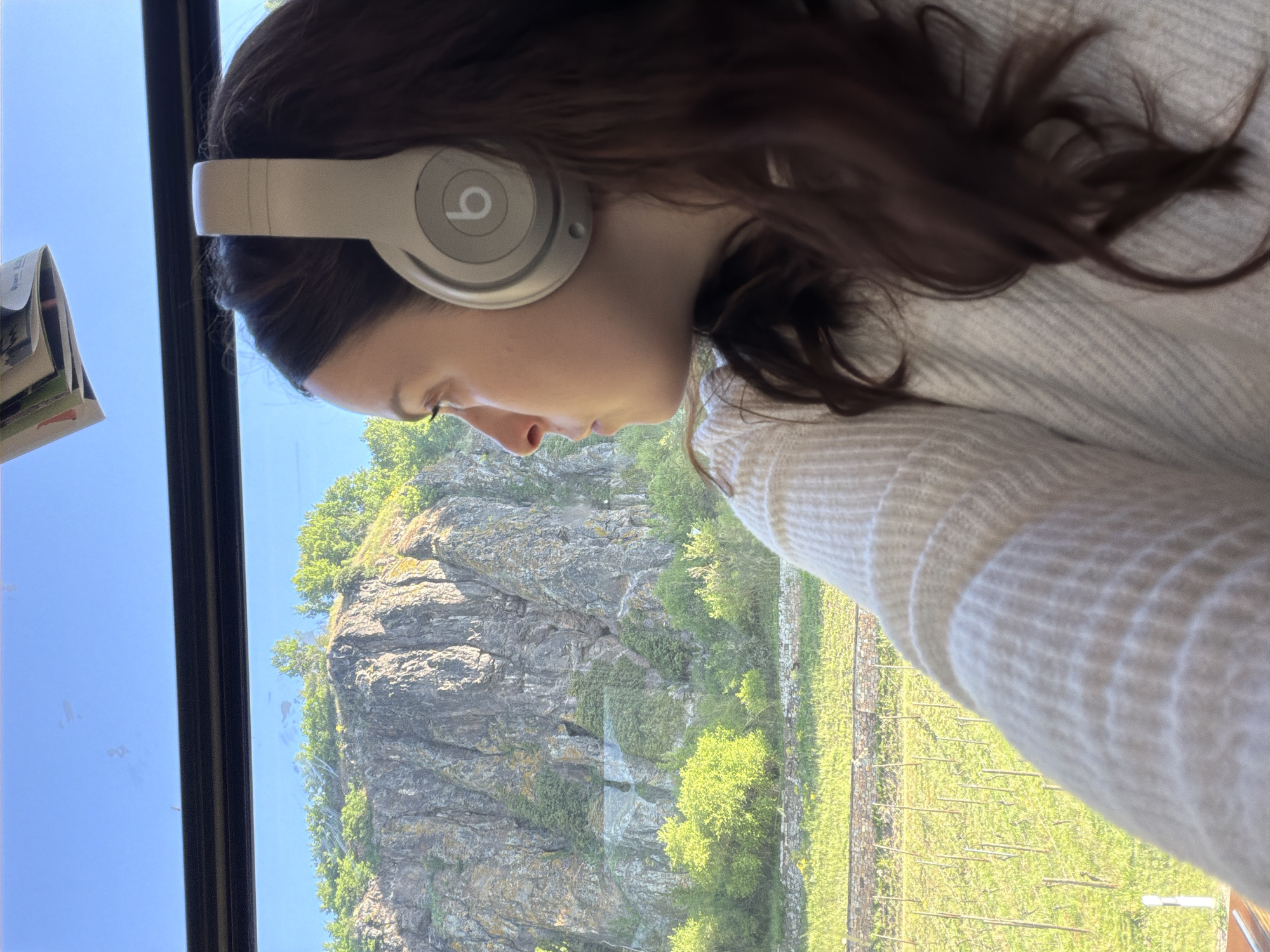 The image shows a young woman with brown hair wearing Beats headphones, looking out a window. The view outside the window shows a rocky cliff face with vegetation and a vineyard. Based on the vineyard and cliff face, the location is likely in Hüffelsheim, Germany. The woman is wearing a light-colored sweater. The window is black framed and likely the window of a train or bus.