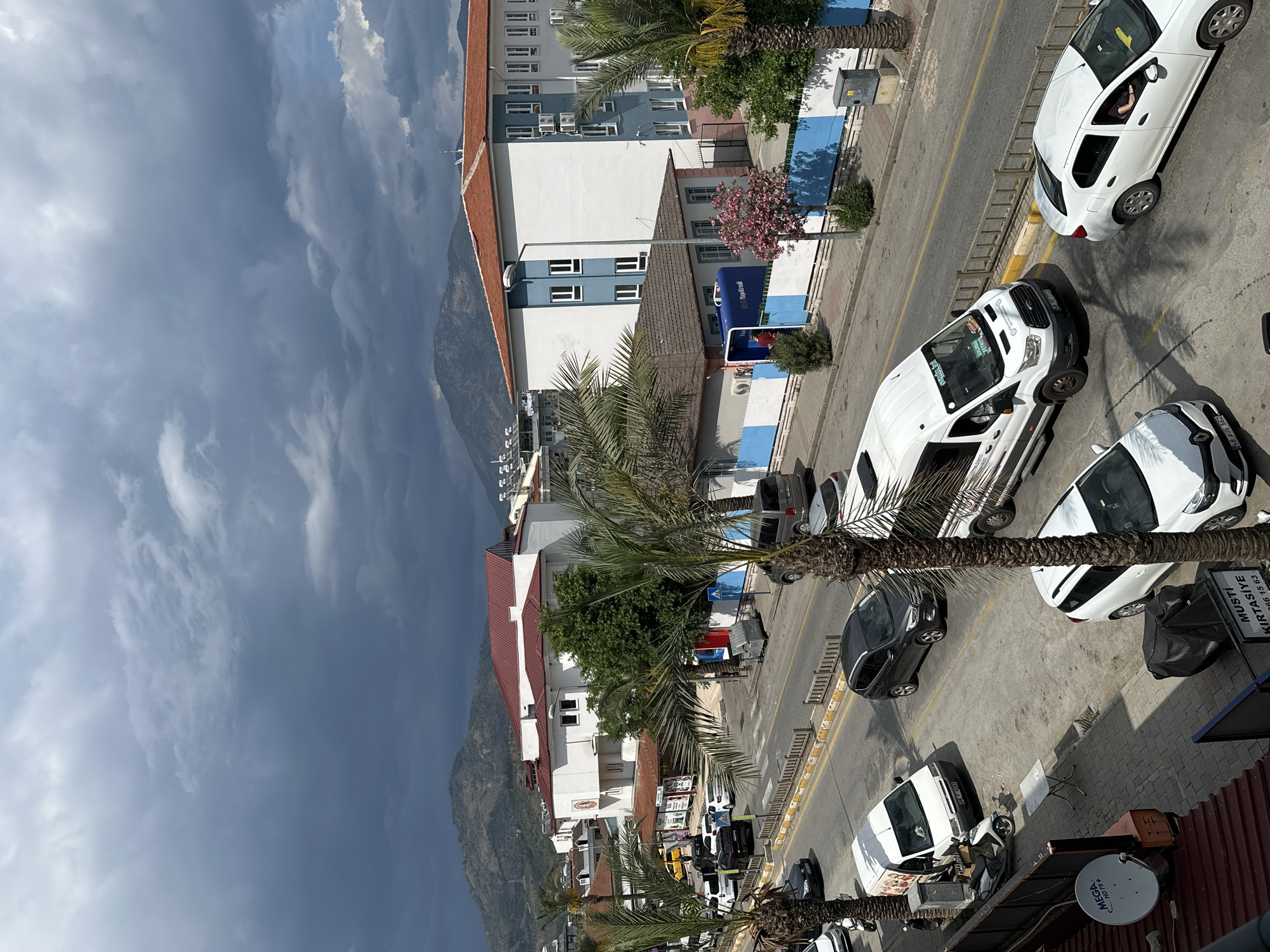 This is an eye-level, landscape-oriented shot of a street in the city of Fethiye, Turkey. The sky is filled with dark, cloudy conditions. A mountain range is visible between the clouds and a series of buildings.
Buildings line the street, showcasing a mix of architectural styles with white, red, and blue color schemes. Palm trees punctuate the skyline, their leaves adding a tropical touch to the urban landscape.
Cars are parked along the side of the road, including white cars and a white van. The street itself is marked by yellow lines.
The foreground includes a partial view of a rusty roof, a satellite dish, and a sign. The sign appears to advertise "MUSTİ KIRTASİYE" and provides a phone number.