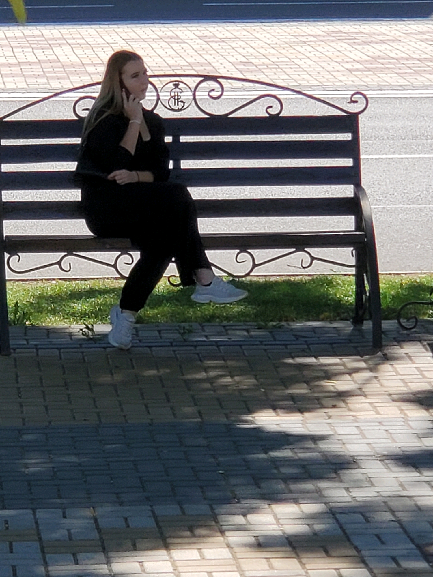 In Tiraspol, Moldova, a young woman with blonde hair is seated on a dark metal park bench, engrossed in a phone conversation. She is dressed in black clothing, including pants and a long-sleeved top, and wears white athletic shoes. The bench is situated on a paved walkway with a tiled pattern, adjacent to a strip of green grass. Behind the bench, a road is visible, with a patterned sidewalk in the background. The scene is bathed in sunlight, casting shadows from the surrounding trees onto the walkway.