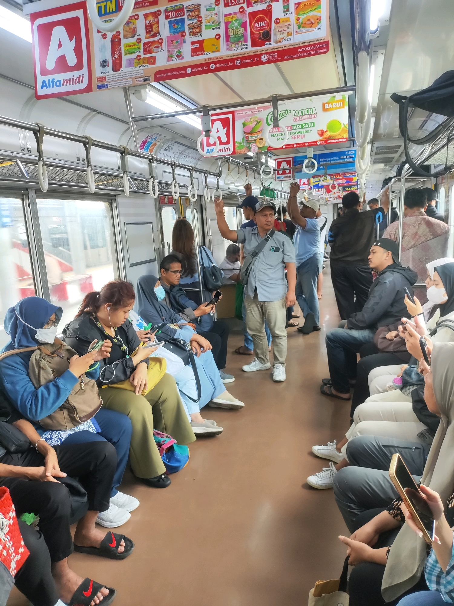 The image depicts the interior of a public train carriage in East Jakarta, Indonesia, with several passengers seated and standing. The carriage is well-lit, with advertising posters visible on the ceiling and upper walls. Passengers, dressed in casual attire, are seen using mobile phones, looking out the windows, or holding onto handrails. The flooring is a light brown color, and white hand straps are hung from the ceiling. Some passengers are wearing face masks, indicating a health-conscious environment. The overall scene conveys a sense of daily commuting within the bustling city.