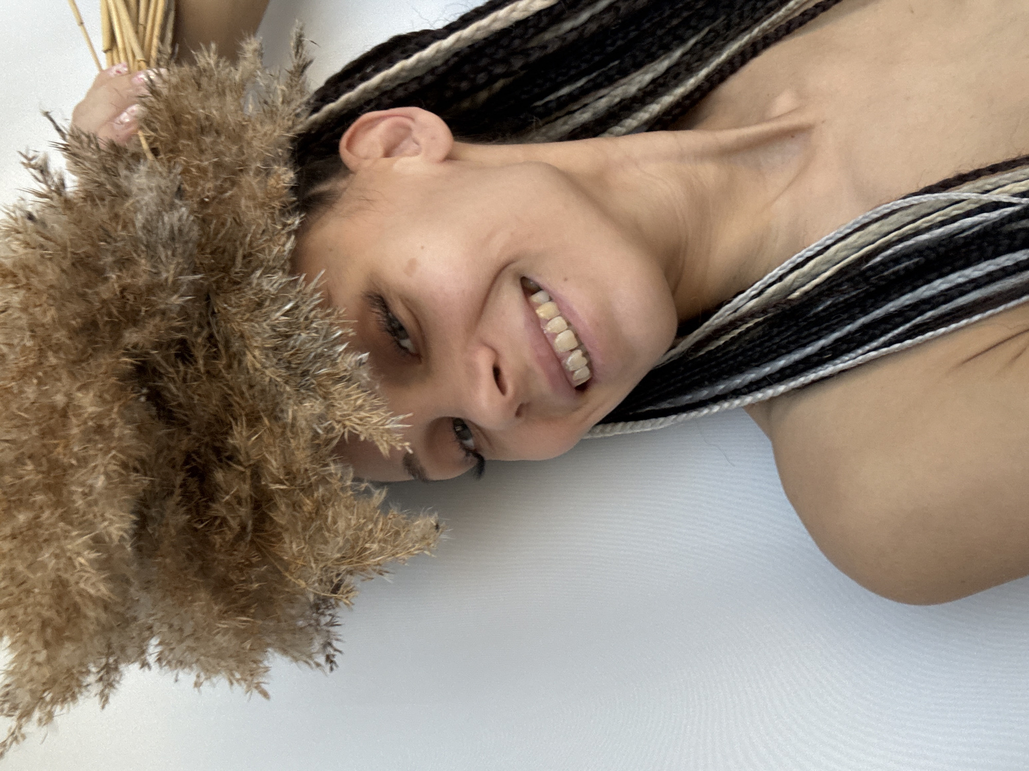 A woman with light brown skin and braided black and white hair is smiling in the image. She is holding a bouquet of dried grass or reeds. The background is a solid white surface. The location is unknown.