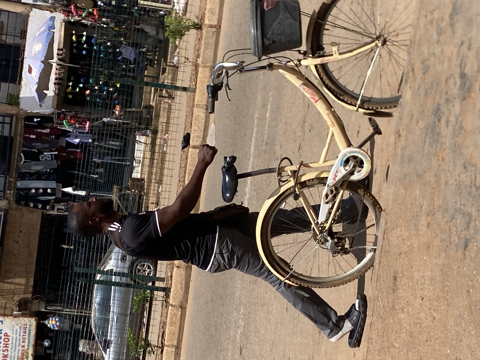 The image captures a man pushing a bicycle on a sunlit street in what appears to be a market area, with the location context being Bukuru, Nigeria. The man, who has a dark complexion and short hair, is dressed in a black t-shirt with white stripes on the sleeves, dark grey trousers, and black and white athletic shoes. He is positioned to the right of a cream-colored utility bicycle, holding its handlebars with his left hand. The bicycle is equipped with a front basket and has fenders over its wheels.
In the background, on the left side of the frame, are several market stalls and shops. These stalls feature metal racks displaying a variety of goods, including clothing and shoes. One stall is partially shaded by a light blue umbrella. A silver car is visible parked behind a metal barrier or fence. The ground is a mix of paved and dirt surfaces, and the clear sunlight indicates it is daytime, with shadows cast by the subjects.
A partially visible sign in the bottom left corner displays text that reads "WORKSHOP", "SALES & RETAILS", and a phone number: "08064588519". The scene suggests a typical busy street with commercial activity.