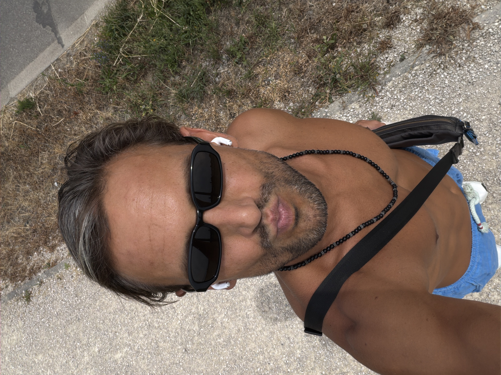 A man is taking a selfie outdoors. He is shirtless, wearing blue shorts, sunglasses, and wireless earbuds. A black crossbody bag is slung over his shoulder. He is standing on a textured surface, possibly a sidewalk or path, with dry grass and a road visible in the background. The overall scene suggests a casual, outdoor setting. The location is Veigy-Foncenex, France.
