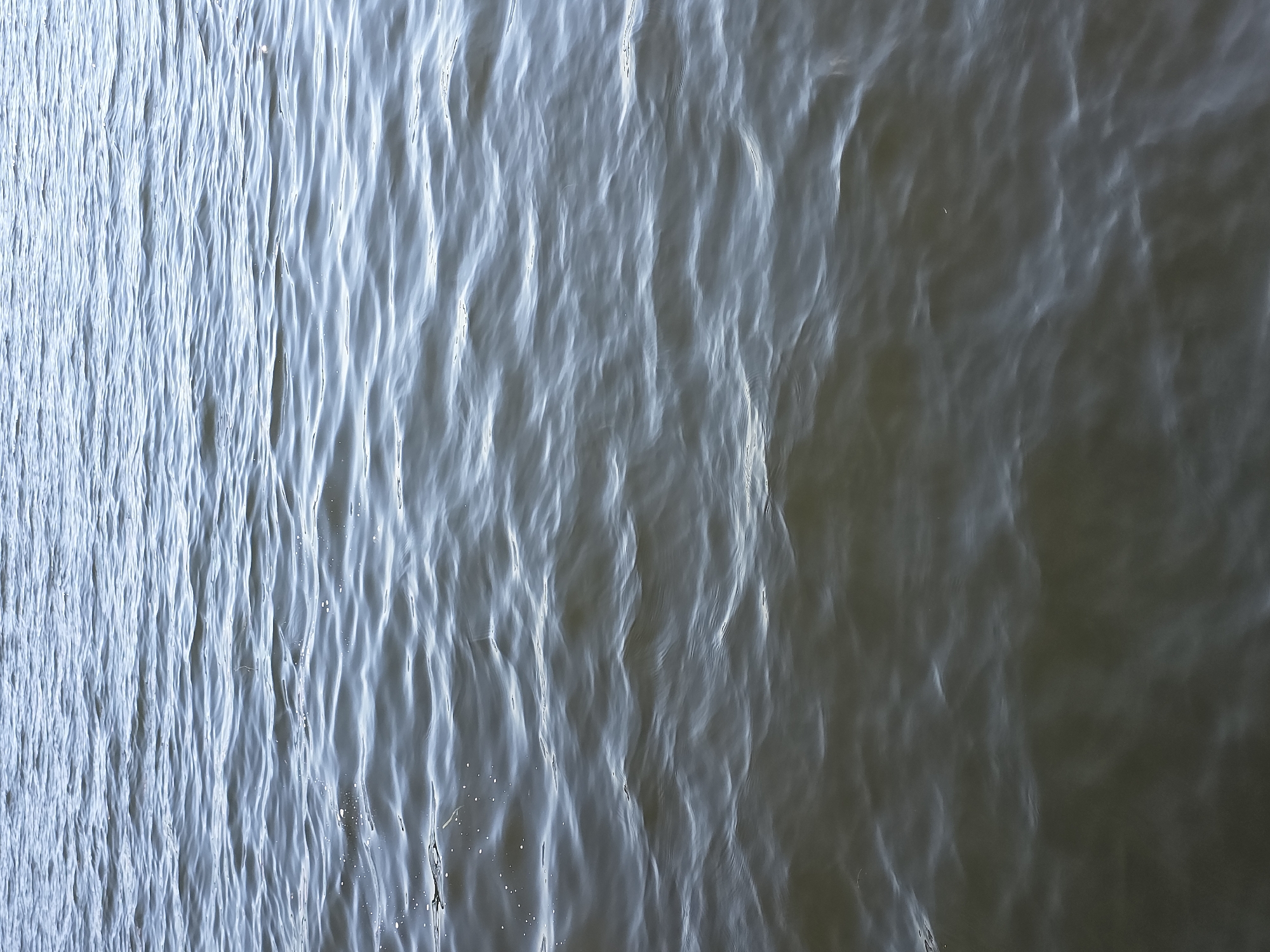 A close-up, head-on shot captures the surface of a body of water, likely Lake Chebarkul in Chebarkul, Russia, under daylight. The water exhibits gentle, undulating waves and ripples, creating a textured pattern across the frame. The coloration is a muted mix of deep blues and grays, with lighter reflections on the crests of the waves, suggesting an overcast or twilight sky. Some small, bright specks, possibly foam or debris, are visible on the surface. The image is devoid of any human presence or distinct objects, focusing solely on the natural movement and appearance of the water.