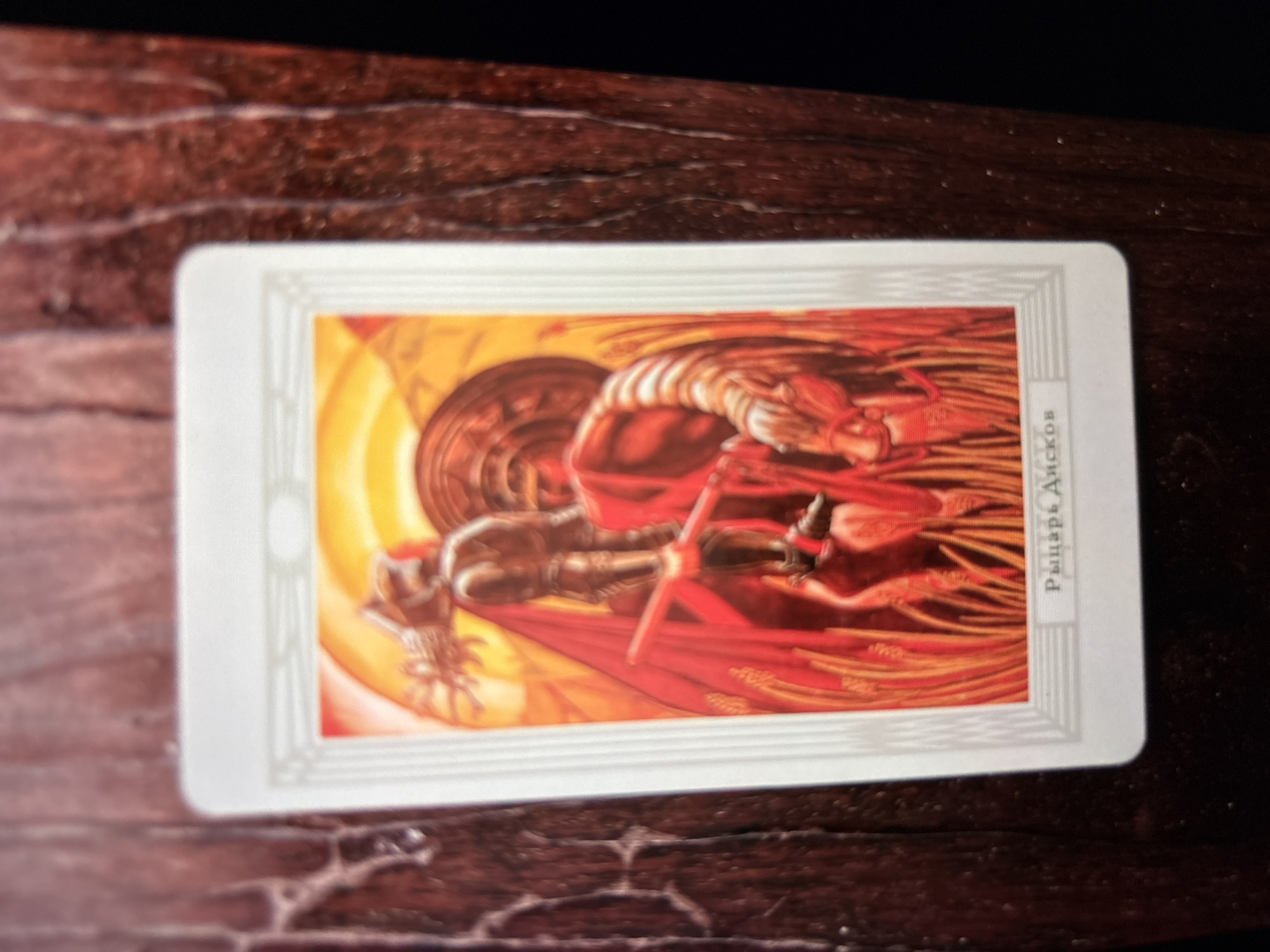 The image is of a tarot card, "Knight of Disks." The card features an illustration of a knight in armor riding a horse across a field. The card has a white border, and the Russian text "Рыцарь дисков" is vertically oriented on the right side of the card. The background is blurred, with a dark wood surface visible. It is not possible to confirm the location of the image to be in the city of Ембаево, Russia with certainty.