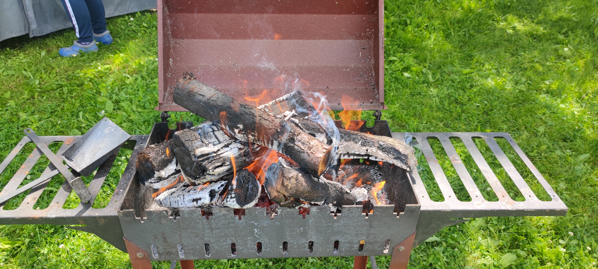 In the city of Пацейкаўскі сельскі Савет, Belarus, an outdoor scene unfolds with a portable metal grill as the central subject. The grill, a dark gray color with a reddish-brown lid, is positioned on a vibrant green grassy lawn. Inside the grill, a fire burns intensely, with logs and embers glowing orange and red, producing wisps of white smoke that curl upwards.
To the left of the grill, a person's legs are visible, clad in dark blue pants with white stripes and bright blue protective footwear. They are standing on the grass, suggesting a casual outdoor activity like a picnic or barbecue.
On the left side of the grill, a metal tool, possibly a shovel or poker, rests on a grated surface. This surface appears to be a shelf or extension of the grill. The grass surrounding the grill is short and well-maintained, with some patches of clover. The lighting suggests it is daytime, with dappled sunlight filtering through unseen trees, casting soft shadows on the grass. The overall impression is one of a preparation for or ongoing outdoor cooking event in a park or backyard setting.