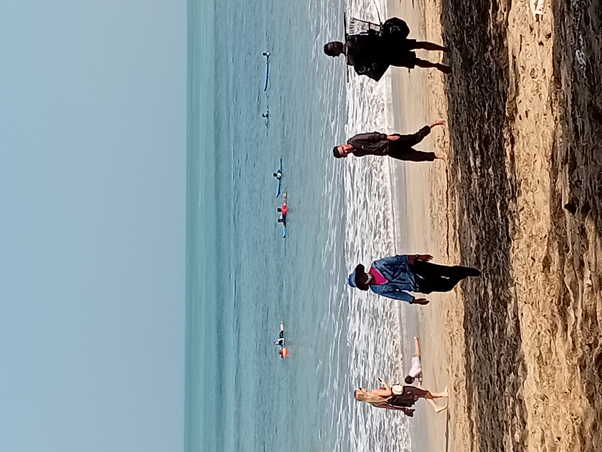 This is a wide-angle, eye-level shot taken on a beach in Kuta, Indonesia. The ocean, a turquoise blue, occupies the upper half of the frame, with a clear, pale blue sky above it. Gentle waves break on the shore, creating a white foam line. Several people are visible on surfboards in the water, appearing to be beginners or waiting for waves.
On the sandy beach, in the foreground and mid-ground, there are several individuals. On the left, a blonde woman in a black dress and a yellow purse is looking at her phone while walking. A person in a white shirt and light-colored shorts is lying face down in the sand near her. Moving towards the center, a person wearing a blue denim jacket, a pink top, dark pants, and a blue hat walks towards the viewer. To their right, a young man in a dark grey hoodie and black pants walks away from the viewer. Further to the right, another person in dark clothing, carrying a large black bag and what appears to be a rack or stand with multiple items, is walking away from the viewer. The sand is light brown and textured, with some shadows indicating it's a sunny day.