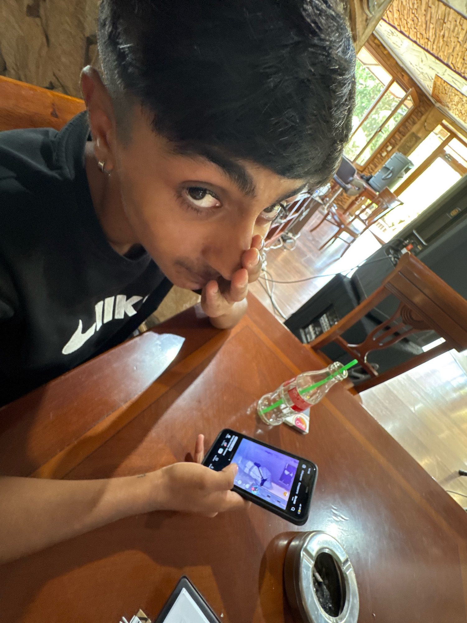 The image shows a close-up of a young person holding a smartphone on a wooden table. The person is wearing a black sweatshirt with a white logo. A glass bottle with a green straw and a Coca-Cola label is on the table, as well as an ashtray. The background suggests an indoor setting with wooden architecture and large windows that allow natural light to enter. The lighting is bright, and the overall scene appears to be a casual dining or gathering space. The location is identified as Sredets, Bulgaria.